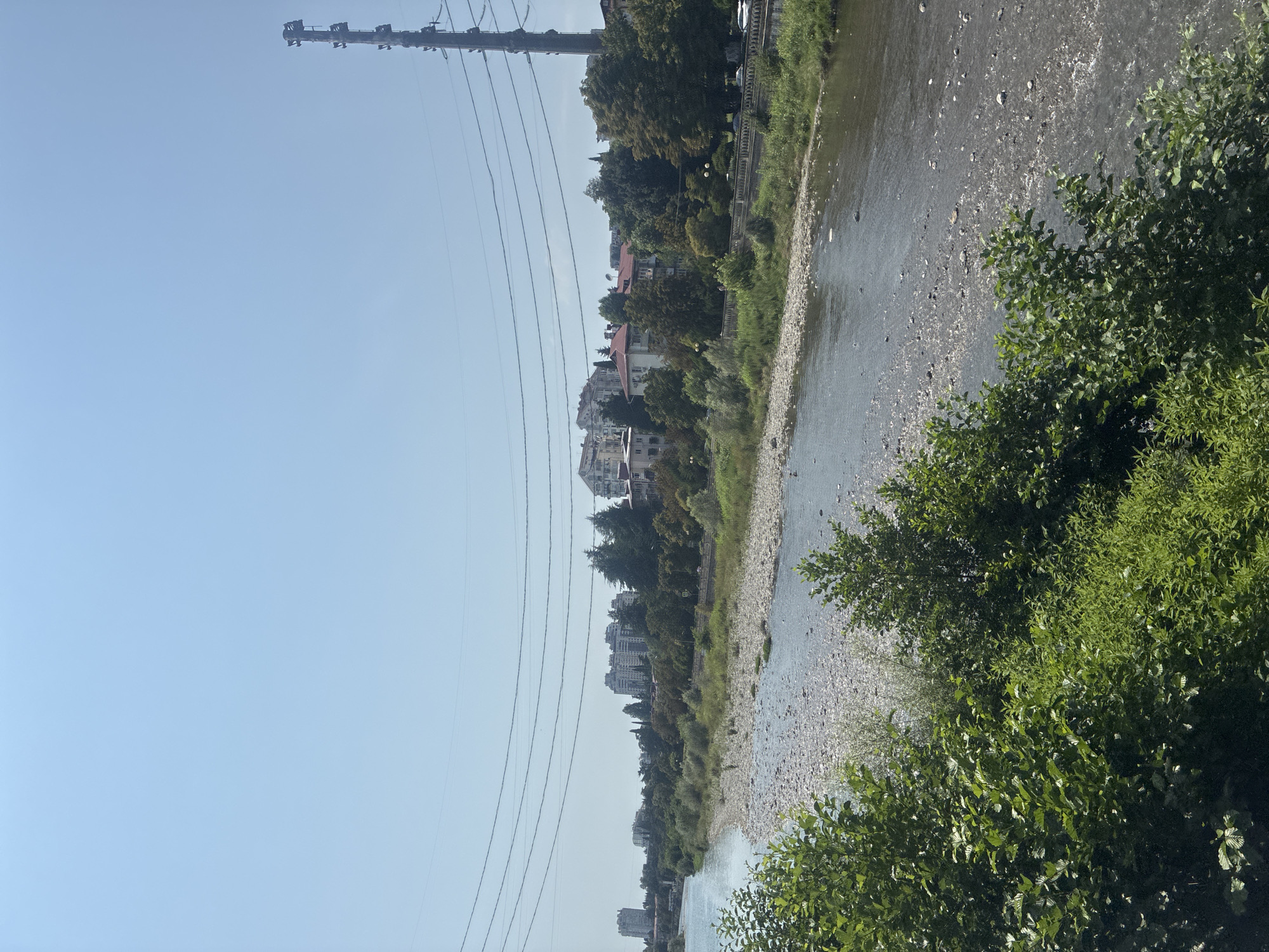 This is a wide, eye-level shot of the Mzymta River flowing through Sochi, Russia. The river is relatively wide with a rocky, shallow riverbed visible in many areas. The water is a murky blue-grey. The foreground is dominated by lush green foliage of trees and bushes. Across the river, on the far bank, there is a well-maintained embankment with a stone wall and lampposts. Behind the embankment, a line of buildings, ranging from mid-rise apartment complexes to smaller houses with red roofs, stretches along the river. Dense green trees are interspersed with the buildings. In the background, a tall, metal telecommunications tower stands to the right of the frame. The sky is clear and blue, with several power lines running horizontally across the upper portion of the image. The overall scene suggests a sunny day in a developed urban area bordering a natural river.