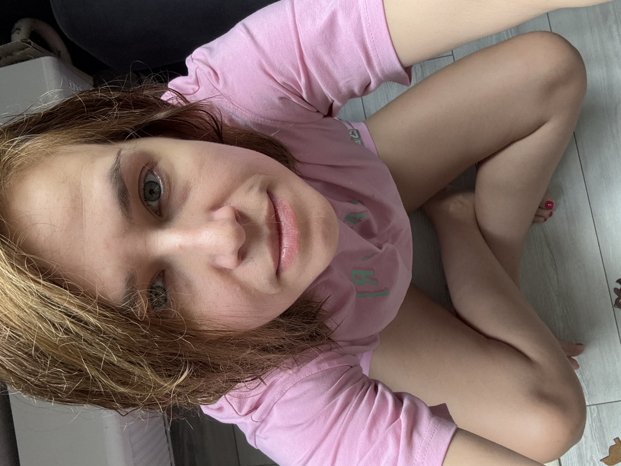 A high-angle, medium shot captures a woman from the chest down, kneeling on a light-colored floor that appears to be tiled. Her legs are bent, with her knees touching the floor and her feet tucked beneath her. She is wearing a short-sleeved, light pink t-shirt with a graphic design on the front. The t-shirt is loose-fitting and extends over her upper thighs. Her hair is light brown and appears damp, falling loosely around her face and shoulders. Her face is turned towards the camera, with her eyes looking directly into the lens. Her skin is pale, and she has a faint smile. The background is out of focus, but hints of dark fabric and a light-colored surface can be seen. The overall setting is indoors, with no specific Moscow landmarks or identifying features of the city visible.