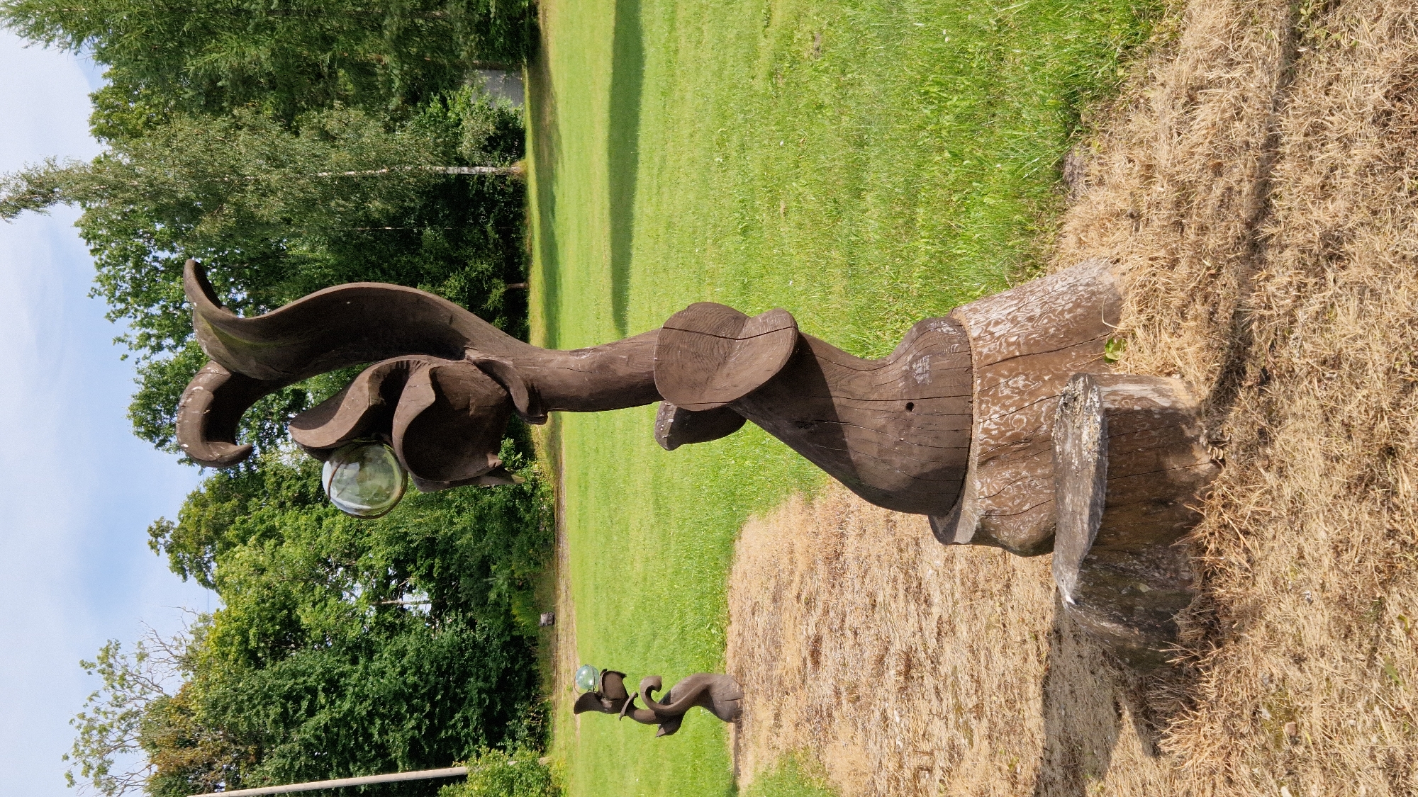 The image depicts a wooden sculpture resembling a stylized plant with a round, transparent glass sphere embedded near its top. The sculpture is dark brown and appears weathered. It is mounted on a thick tree stump. In the background, another similar, smaller sculpture is visible further down a dry, grassy path. The environment is a park or garden with lush green grass, trees, and a bright sky with some clouds. The scene is located in the vicinity of Kazdanga, Latvia, as indicated by the provided context. There are no people visible in the image. The overall impression is one of outdoor art installation in a natural setting.