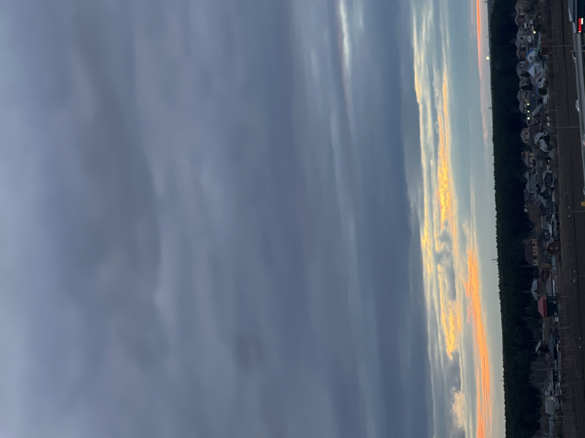 The image captures a twilight scene over Tyumen, Russia, likely during sunset or sunrise. The horizon runs horizontally across the lower third of the frame, with the sky dominating the upper two-thirds. Large, dark grey and bluish clouds fill most of the sky. Towards the horizon, these clouds are dramatically illuminated by vibrant streaks of bright orange and yellow light, suggesting the sun is just below the visible line.
Below the illuminated clouds, a dense, dark strip of trees, likely a forest, stretches across the mid-ground. Further towards the bottom edge of the image, a residential area or settlement is visible, featuring numerous houses with various rooflines. Several small, bright points of light are discernible within the settlement, indicating artificial illumination from windows or streetlights. Along the far bottom edge, parallel to the settlement, there is a dark strip of infrastructure, possibly a road or railway line, with some indistinct shapes that could be vehicles or other structures.
The scene conveys a sense of calm, with the warm hues on the horizon contrasting against the dark sky and foreground elements. No people or visible text are present in the image.