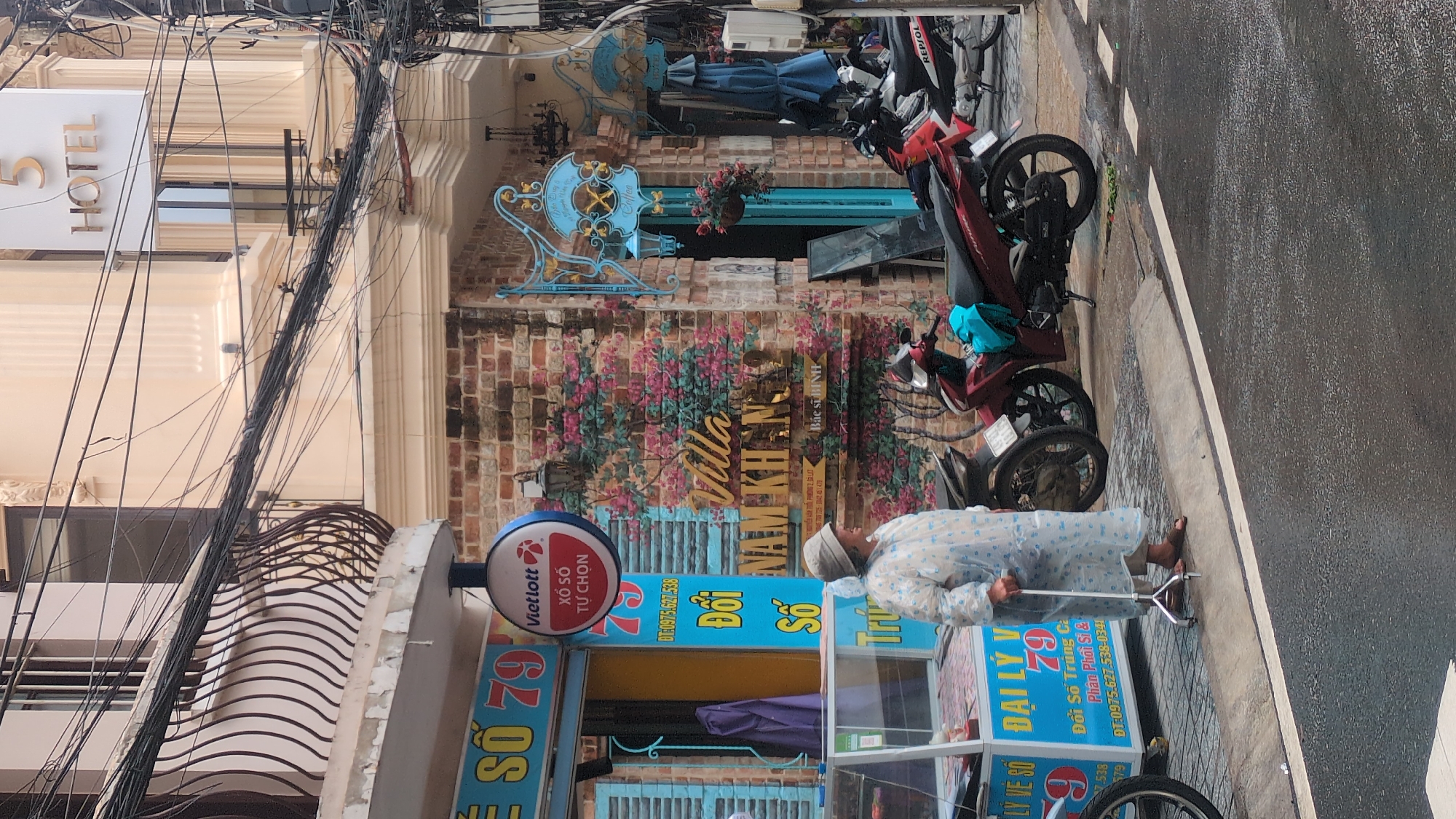 The image depicts a street scene in Da Lat, Vietnam, with a focus on the facade of a building and the area in front of it. On the left, a lottery shop with "Số 79" signage is visible, featuring blue shutters and a display case. A woman in a light-colored raincoat and hat, leaning on a cane, stands on the sidewalk. To the right of the lottery shop, a brick wall is decorated with a floral mural. Above the mural, a sign reads "Villa Nam Khanh 2" in gold lettering. Further to the right, a blue sign with script and a coffee cup motif is mounted on the wall, suggesting a cafe or establishment. A building with a "5 HOTEL" sign is visible in the background. Several motorbikes are parked along the street, some in the foreground and others further back. The street appears wet, indicating recent rain. The overall scene suggests a bustling urban environment with various commercial establishments.