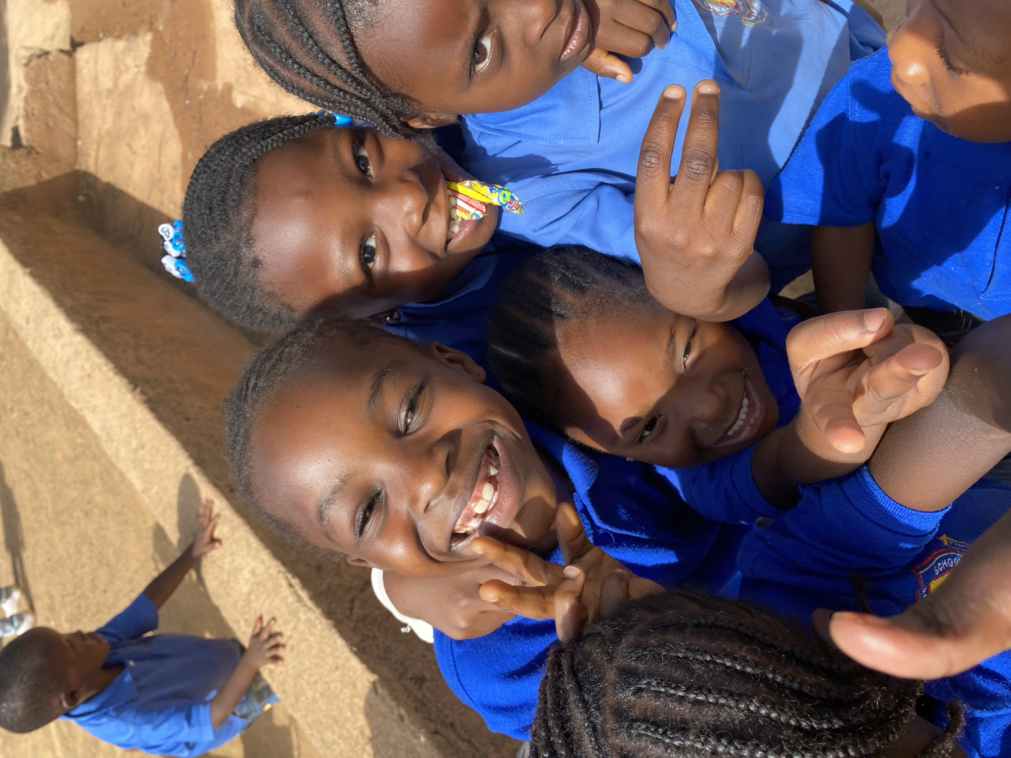 A group of five African children in blue school uniforms are posing for a photo together. The photo is taken from a slightly elevated, downward-looking perspective. The children are huddled closely together, their faces directed towards the camera. One of the children is holding up two fingers in a peace sign. The environment appears to be outdoors, potentially a schoolyard, with a sunlit wall visible in the background on the left side of the frame. Another child in a blue shirt is visible standing near the wall. Based on the information provided, the location is Gura Top, Nigeria.