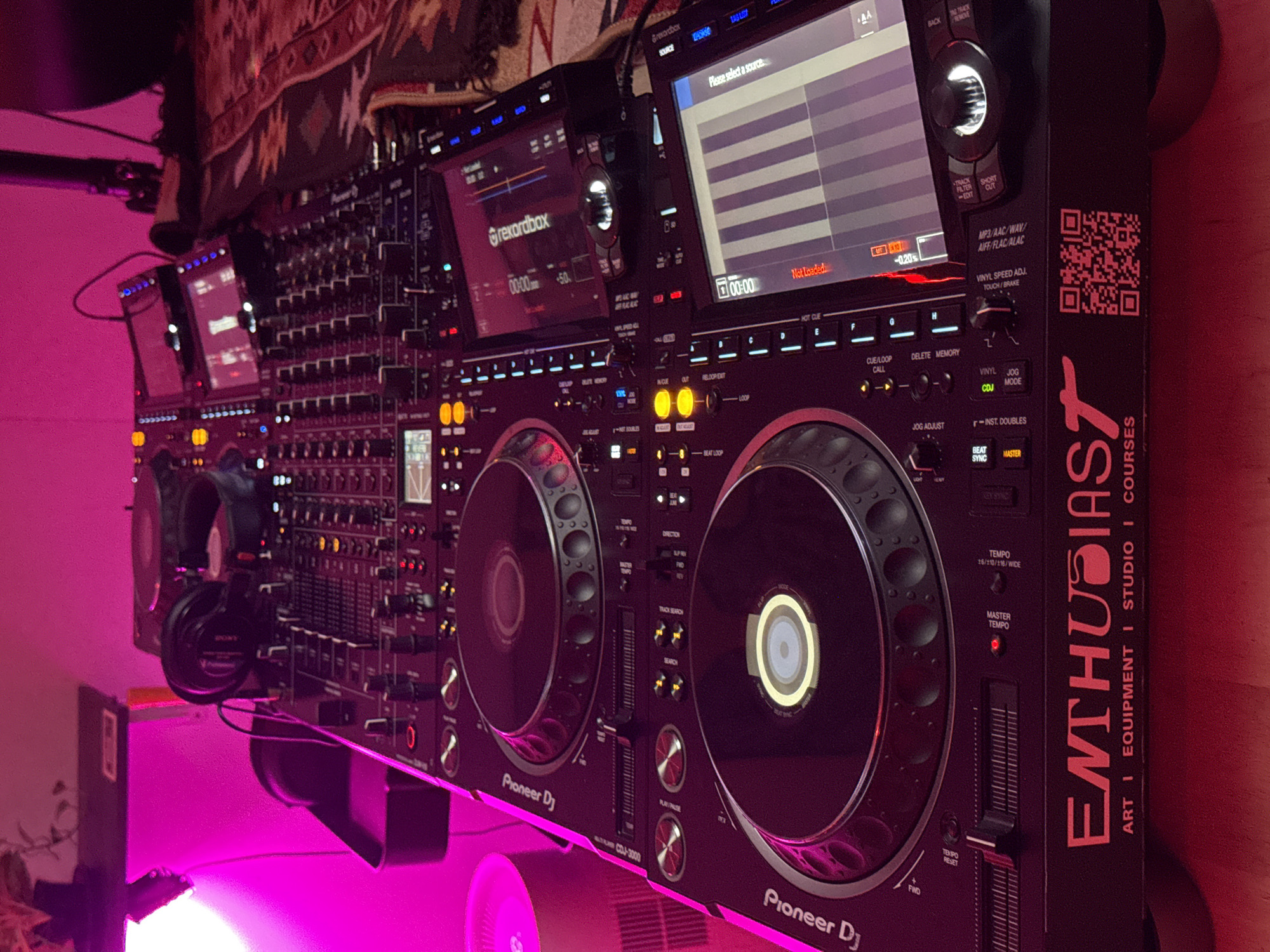 A top-down, angled shot captures a professional DJ setup in a dimly lit room with vibrant pink lighting. The focus is on a complex Pioneer DJ system, including two turntables and a mixer with multiple buttons, sliders, and screens. Two black headphones rest on top of the mixer. The DJ equipment is positioned on a wooden surface, and a patterned rug is visible in the background on the right. A plant is partially visible in the upper left corner. Text on the DJ system includes "Pioneer DJ" and "Enthusiast ART I EQUIPMENT I STUDIO I COURSES". A QR code is also present. The overall impression is of a prepared space for a DJ performance or practice session.
