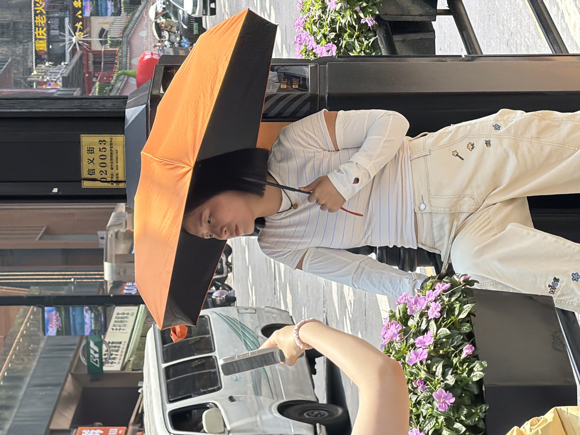 In Yuzhong District, China, a woman is holding an orange umbrella over her head, shielding herself from the sun. She is wearing a white long-sleeved shirt with blue stripes and detached white sleeves, and light-colored pants decorated with small cartoon patches. She is sitting on a ledge next to a planter filled with pink flowers. To her left, another person is holding up a smartphone, appearing to take a photo or video of her. In the background, a white van is parked on the street, and further back, buildings with signage and other pedestrians are visible. A sign on a pole near the woman has Chinese characters and a number "020053".