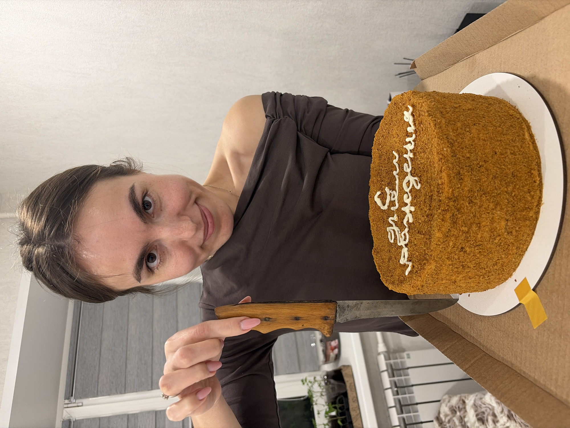 A person with brown hair pulled back is holding a knife next to a cake. The cake is on a white plate inside a cardboard box. White frosting is written on the cake. The person is wearing a brown top and has manicured fingernails. A window with closed blinds is visible in the background. The location is Sarapul, Russia.