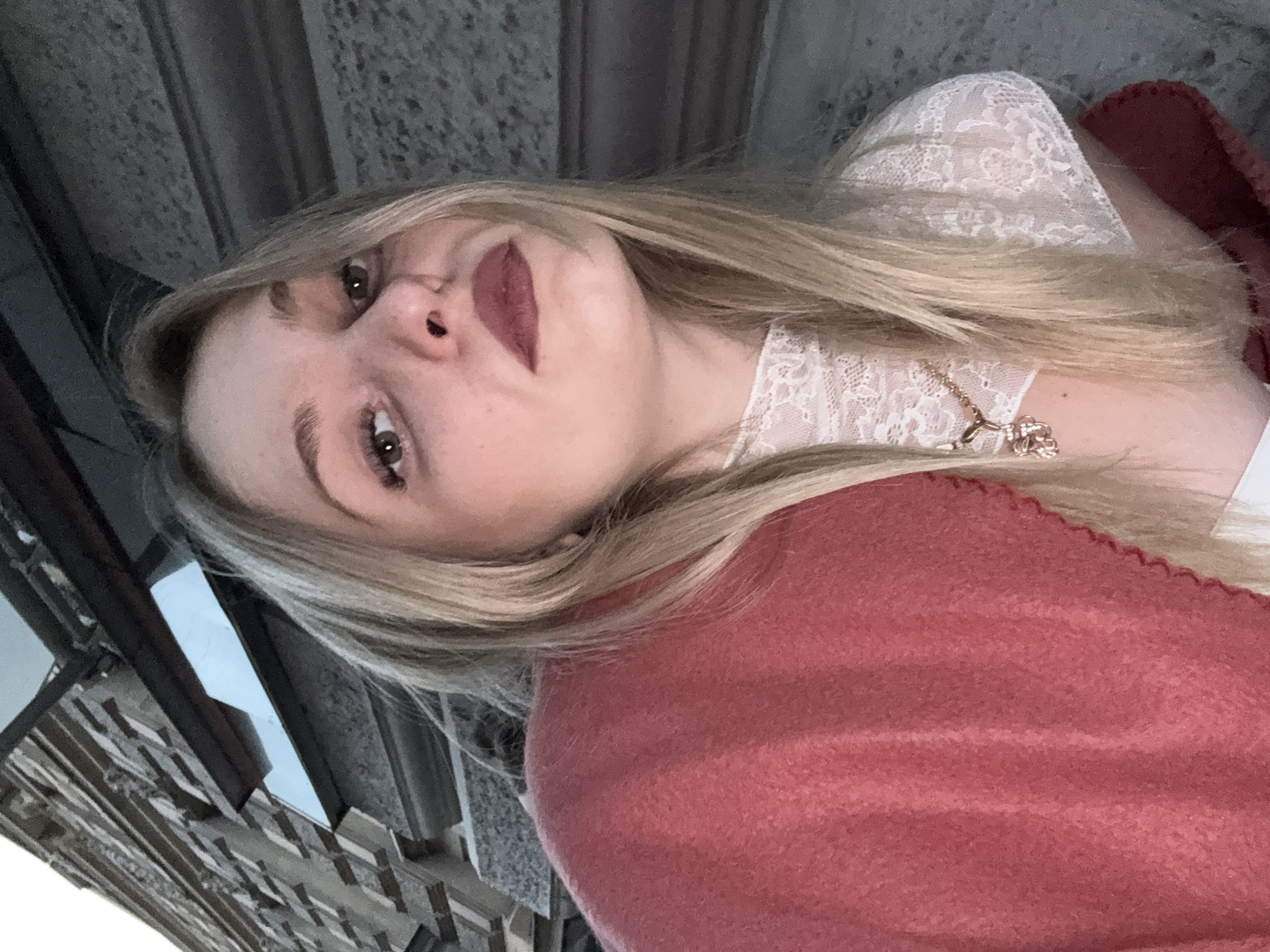 An outdoor, eye-level, medium shot shows a young woman with blonde hair, wearing a red blanket, a white lace top, and a gold necklace. She is leaning into the frame from the left, with her face turned toward the camera. Her eyes are open and she has dark lipstick on. Behind her, there is a stone building and a dark awning. The lighting is low, suggesting it is evening or nighttime.