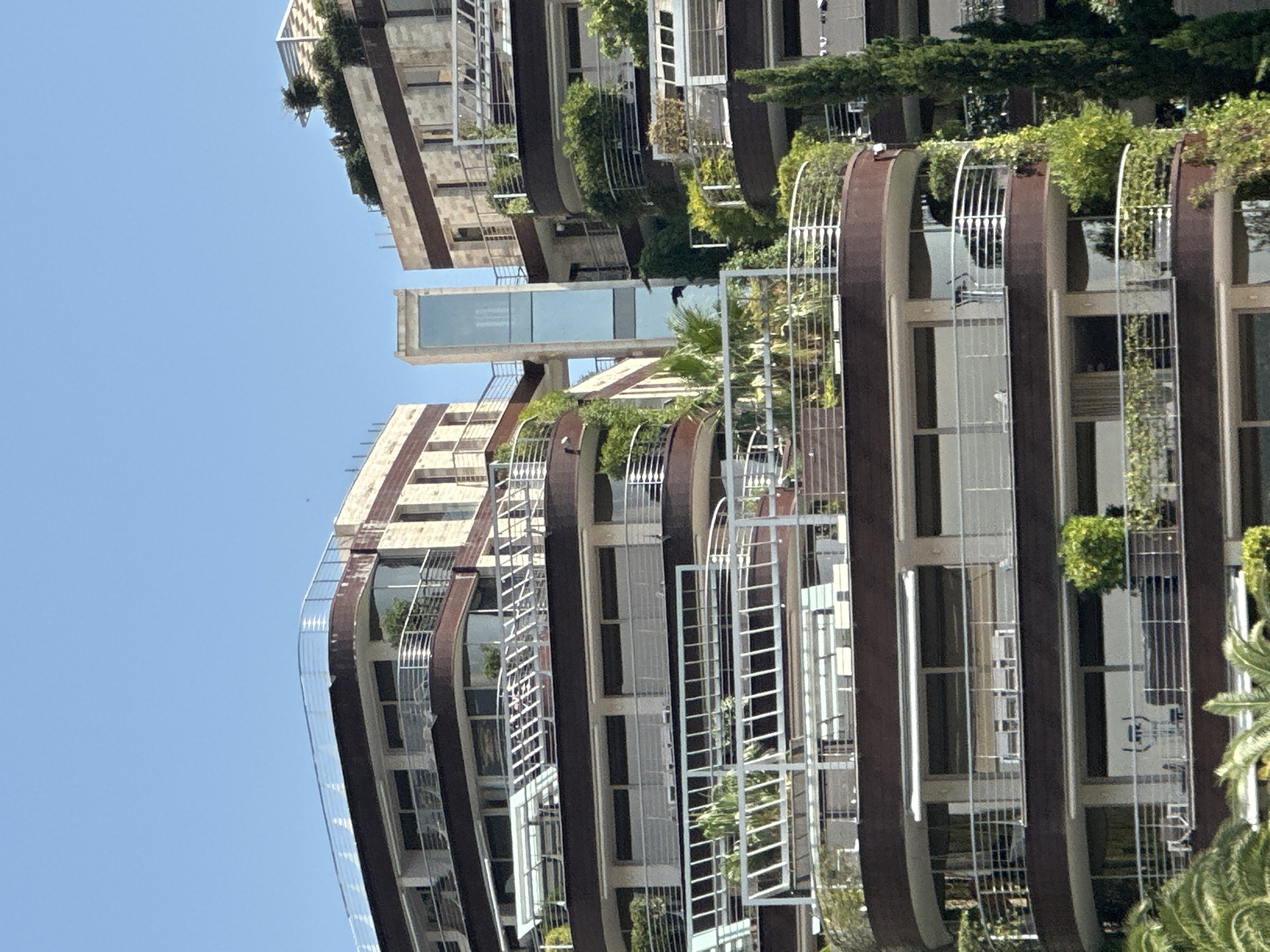 This media file captures a close-up, vertical shot of a modern, multi-story building with a unique architectural design. The building features curved balconies with metal railings, interspersed with sections of windows and what appear to be balconies adorned with lush greenery. The facade is a mix of light-colored stone or concrete and darker, possibly metallic, vertical accents.
The building is bathed in bright sunlight, suggesting it is daytime, and the clear blue sky in the background indicates fair weather. The image is oriented vertically, making it appear as if the building is extending upwards from the bottom or downwards from the top, depending on the viewer's perspective.
There are no people visible in the frame. The primary focus is the building itself, its structure, and the integration of vegetation, creating a sense of modern living with a touch of nature. The reflection in the windows is subtle, showing hints of greenery and possibly other structures, but no distinct details.
A prominent horizontal element, a glass-enclosed walkway or bridge, is visible partway up the building, connecting two sections. The overall impression is one of contemporary architecture, potentially a residential building or hotel, given the balcony designs and the presence of greenery.
The location context provided is Budva, Montenegro.