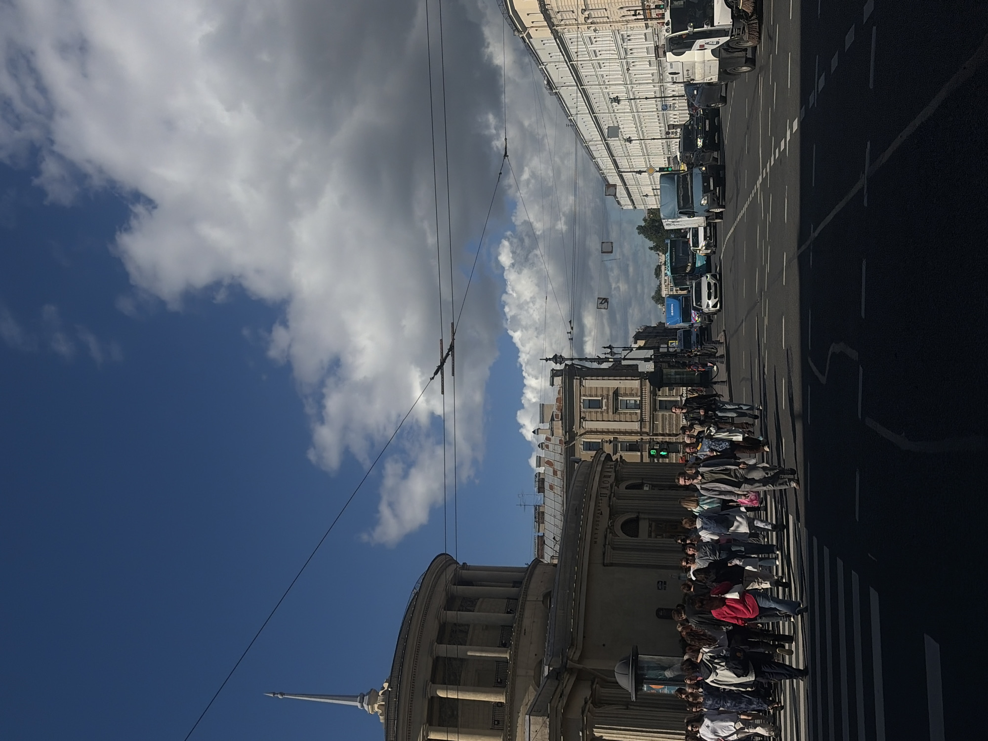 The image depicts a street scene in Saint Petersburg, Russia, with a focus on a group of people crossing a street at a pedestrian crossing. The scene is captured from a low angle, looking upwards.
The subjects are a large group of people, mostly young adults, waiting to cross or in the process of crossing a wide street. They are dressed in casual clothing, suggesting a warm, possibly sunny day. Some are looking at their phones, while others are chatting or looking forward.
In the background, there are grand, classical-style buildings with ornate facades, characteristic of Saint Petersburg's architecture. The street is paved, with clear white pedestrian crossing lines. Several vehicles are visible in the background, including trucks, buses, and cars, suggesting ongoing traffic. Overhead, there are trolleybus wires, a common sight in Saint Petersburg.
The sky is a mix of bright blue and dramatic, dark clouds, indicating varied weather conditions. The sunlight casts shadows on the road, suggesting it is daytime, likely late morning or afternoon.
The overall mood is one of everyday urban life. The people appear relaxed and unhurried. There are no clear emotional expressions to discern.
Some visible text includes a green traffic light signal, and signs on buildings or vehicles, but the text is mostly illegible due to resolution and angle. One sign on a building has Cyrillic text, partially visible as "ПЛОЩАДЬ", meaning "square".