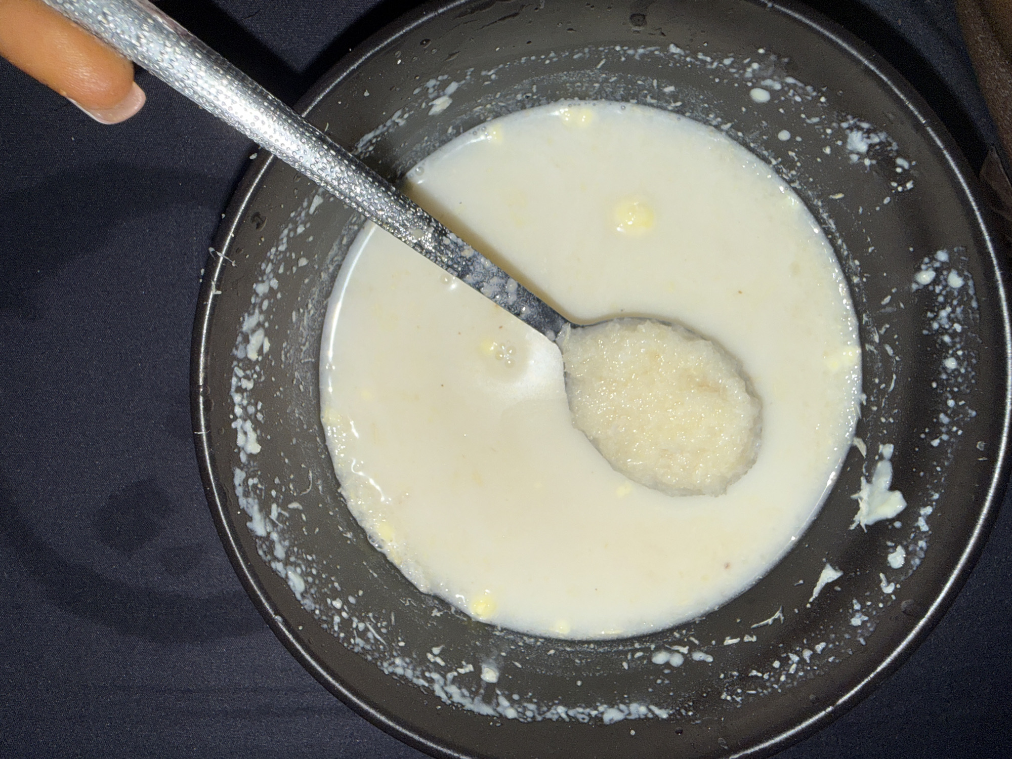 A close-up overhead shot shows a black bowl filled with a creamy, pale liquid and a small mound of grainy, light-colored food in the center, which is being scooped by a textured, metallic spoon. The spoon is positioned at an angle, with a portion of a person's thumb and fingernail visible at the top right, holding the spoon. The rim of the bowl is speckled with dried white droplets, suggesting it has been used or is in the process of being served. The background is a dark, textured fabric, likely a tablecloth. The image does not contain any elements that would suggest a specific location in Abuja, Nigeria.