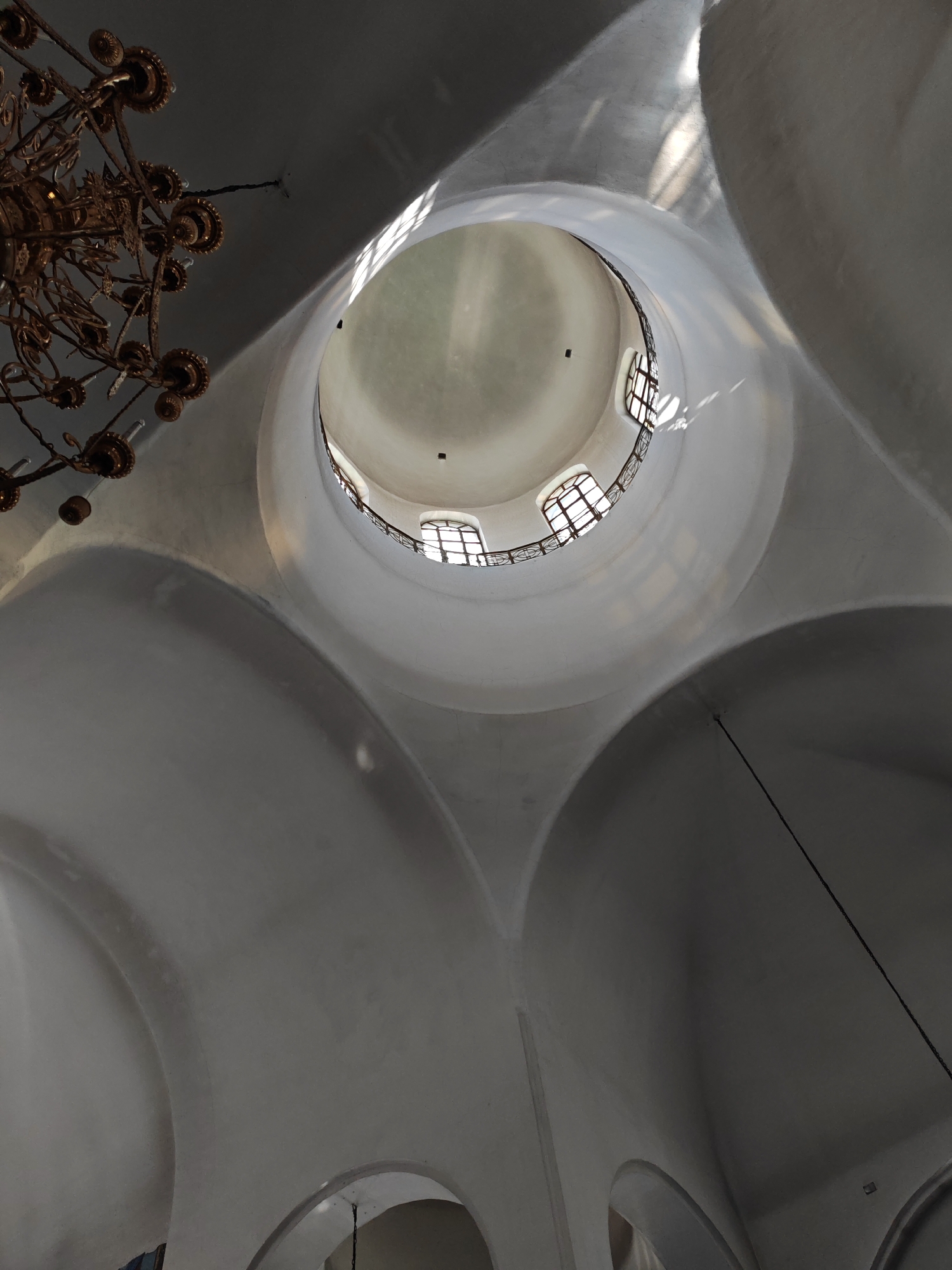 An interior shot captures a grand, possibly church or cathedral, dome with a large circular opening at the apex. Sunlight streams through the opening, illuminating the curved, white plastered interior walls and creating dramatic shadows. A ornate, dark metal chandelier hangs from the upper left of the frame, its intricate branches extending downwards. The dome itself is textured and appears to be constructed of plaster or stucco. Surrounding the central opening, a dark metal railing or balustrade can be seen, with several arched windows visible beyond it. The architecture suggests a classical or baroque style. This image was taken in Yoshkar-Ola, Russia, specifically within the National Art Gallery, which is known for its unique architectural design that resembles a castle.