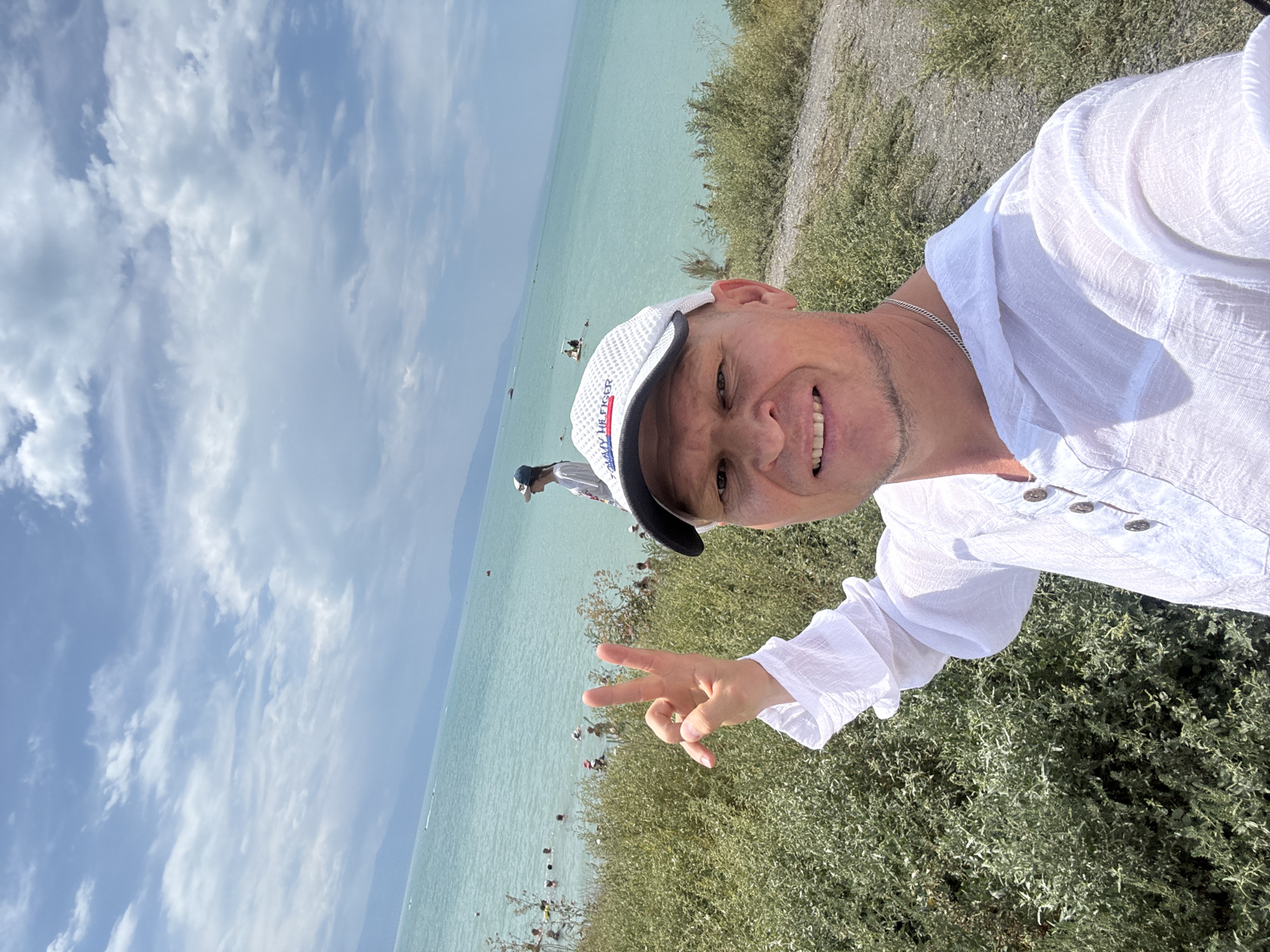 A man is taking a selfie outdoors by a body of water. He is wearing a white shirt with puffy sleeves and a white baseball cap. He is smiling and holding up two fingers in a peace sign. In the background, people are swimming and there are a few boats on the water. The sky is cloudy. The man is standing on a grassy slope overlooking the water. The scene appears to be a lake or large body of water. The location is Urdzhar, Kazakhstan.