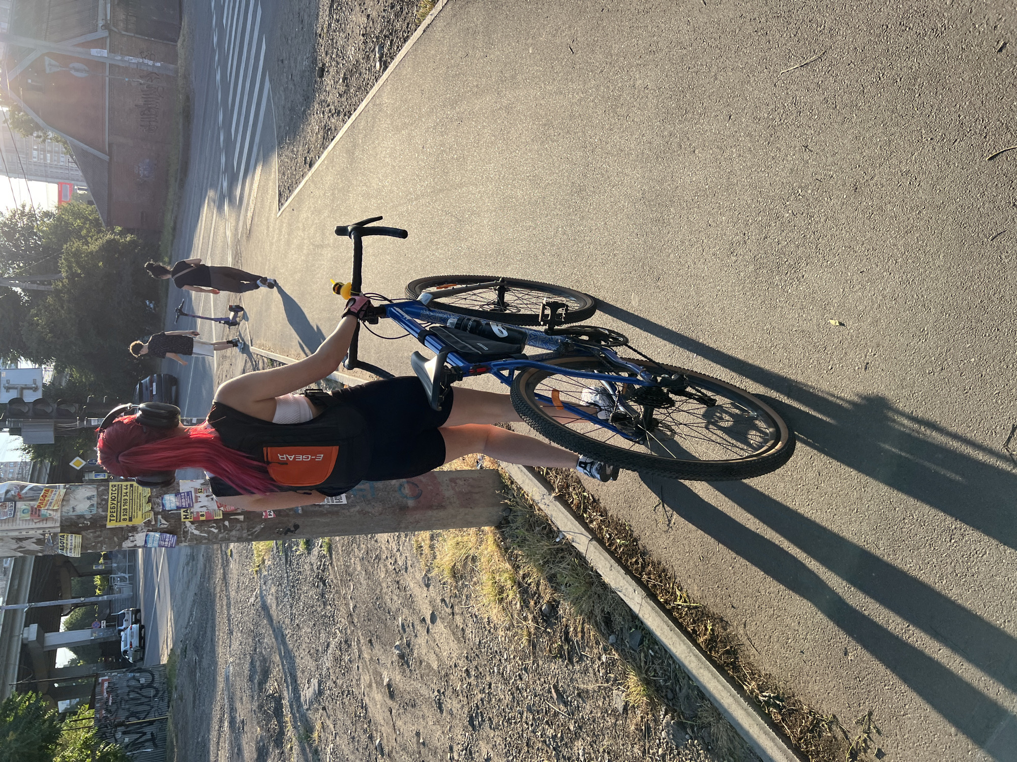 The image depicts a young woman with vibrant pink hair, wearing headphones and a black athletic top with an orange "E-GEAR" pouch, standing next to a blue bicycle. She is wearing black shorts and sneakers. The scene appears to be an outdoor paved area, possibly a bike path or sidewalk, adjacent to a dirt and gravel area. In the background, there are other people on scooters, a crosswalk, buildings, and elevated roads, suggesting an urban environment. The sun is low in the sky, casting long shadows, indicating it is either early morning or late afternoon. The overall mood seems casual and active. Visible text includes "E-GEAR" on the woman's pouch, and various advertisements and Cyrillic text on a pole near her, including what appears to be phone numbers.