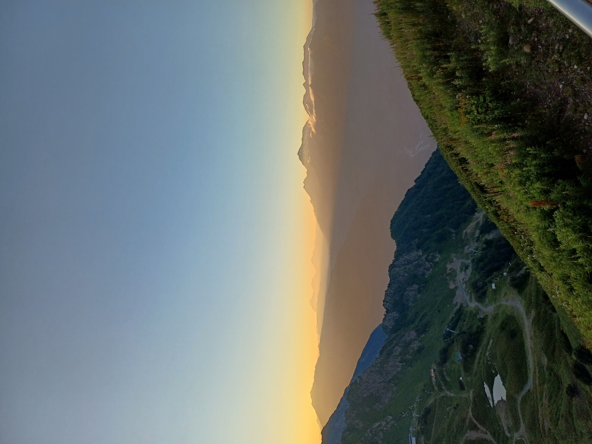 The image depicts a panoramic view of a mountain landscape at what appears to be sunrise or sunset. The sky transitions from a clear light blue at the top to a soft yellow and orange hue near the horizon, illuminating distant mountain ranges. In the foreground, a steep, grassy slope covered in vegetation, including wildflowers, descends to the left. The middle ground features a darker, more rugged mountain slope with visible trails, some small buildings or structures, and at least two small bodies of water, possibly ponds or small lakes. Further into the distance, layers of mountains fade into a hazy, muted brown, with some of the peaks catching the light. The scene is devoid of any visible people or animals. This landscape is consistent with the mountainous terrain surrounding the city of Estosadok, Russia, which is known for its ski resorts and natural beauty.