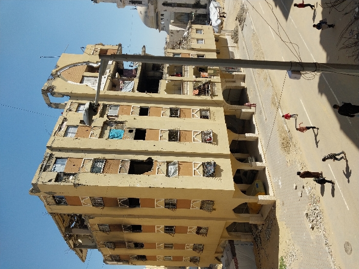 An overhead view captures a street scene in Khan Yunis, Palestinian Territory, dominated by a heavily damaged multi-story residential building under a clear blue sky. The seven-story, light-tan building features dark-brown window trim and an arched ground floor. It exhibits extensive structural damage, including numerous bullet holes across its facade, large sections of missing walls on upper floors, exposed rebar, and scorched areas, particularly on the left side of the third floor. Shattered windows and exposed interior rooms are visible throughout.
On the street below, approximately ten individuals are present. Six children, some wearing red shirts, are dispersed across the asphalt road, running and walking. Two adults are also visible: one male in a brown garment standing near the left sidewalk and another male in dark clothing walking in the immediate foreground with his back to the camera. A yellow vehicle is parked beneath the arches of the damaged building's ground floor.
To the right of the main building, a mosque with a light-colored dome and a partial minaret is visible, appearing worn or damaged. The street features a paved road with a white centerline, flanked by unpaved areas covered in sand, dirt, and debris. A utility pole with a streetlamp and electrical wires extends diagonally across the mid-right of the frame. Shadows are sharply cast on the ground, indicating direct sunlight.