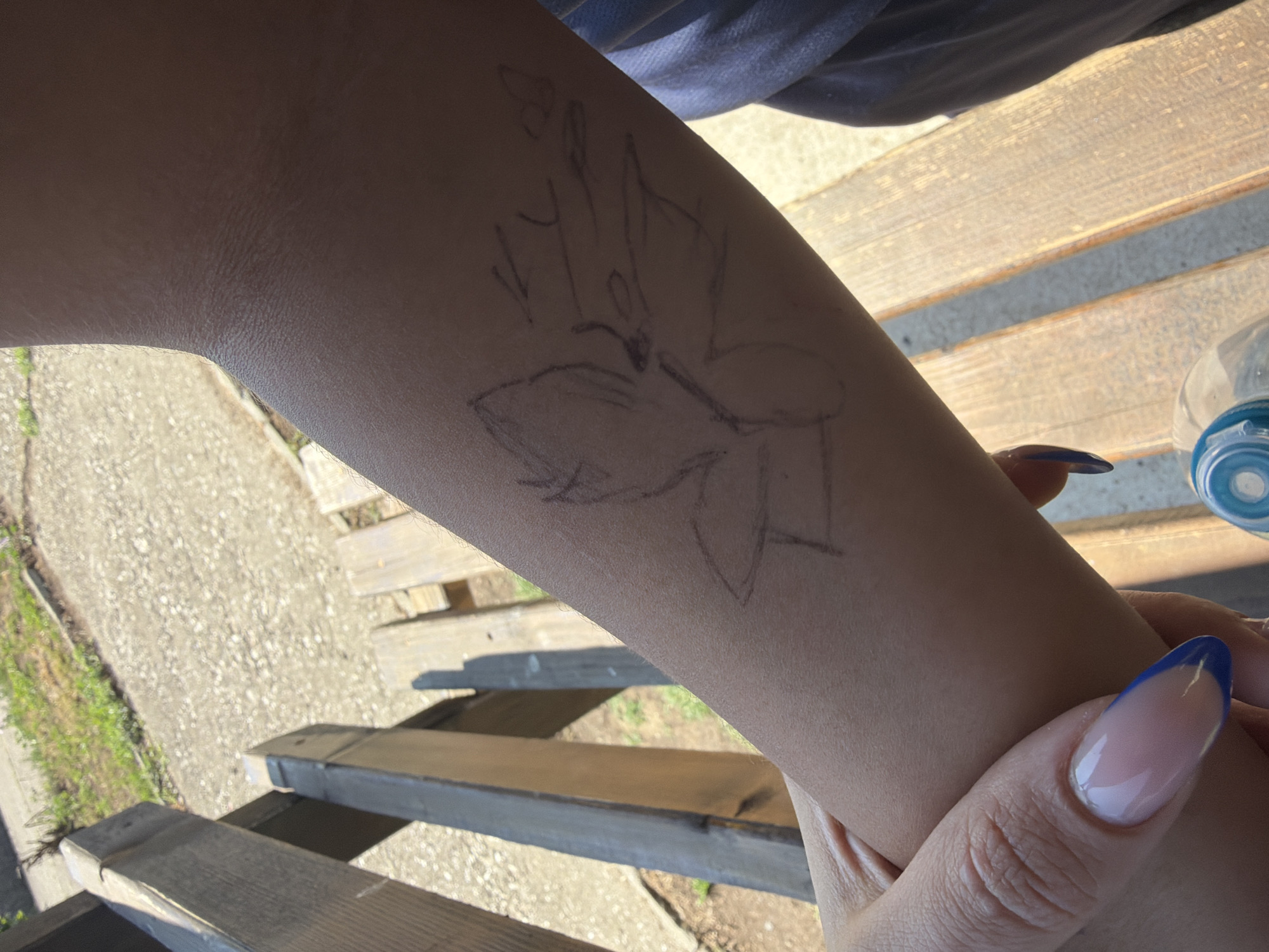The image shows a person's forearm with a rough sketch of a flower or butterfly drawn on it. The person's hand is visible in the foreground with manicured nails, featuring a French tip design with blue accents. They are seated on a wooden bench, with the background showing gravel and some green vegetation, suggesting an outdoor setting. A plastic water bottle is partially visible at the bottom of the frame. The overall lighting indicates it is daytime. No information about Dinkata, Bulgaria, is present in the image.