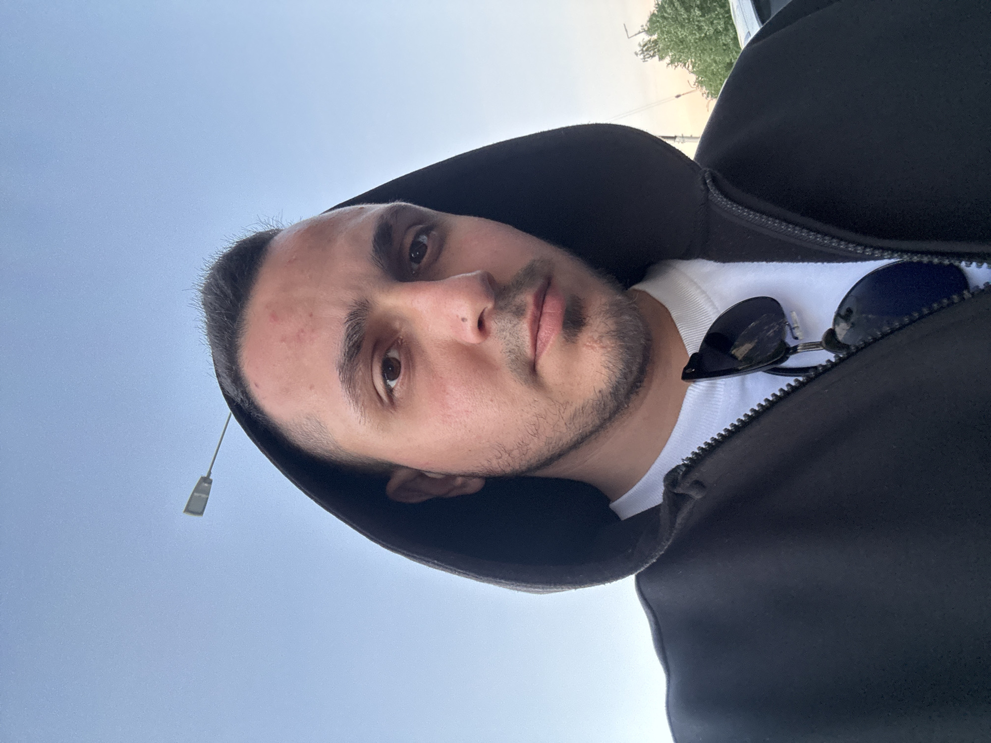 A close-up selfie shows a man with short, dark hair and a light beard. He is wearing a black hooded jacket zipped up over a white t-shirt. A pair of dark sunglasses is clipped to the front of his jacket. The man is looking directly at the camera. The background is a clear sky with a street lamp visible in the distance. The image appears to be taken outdoors during the day.