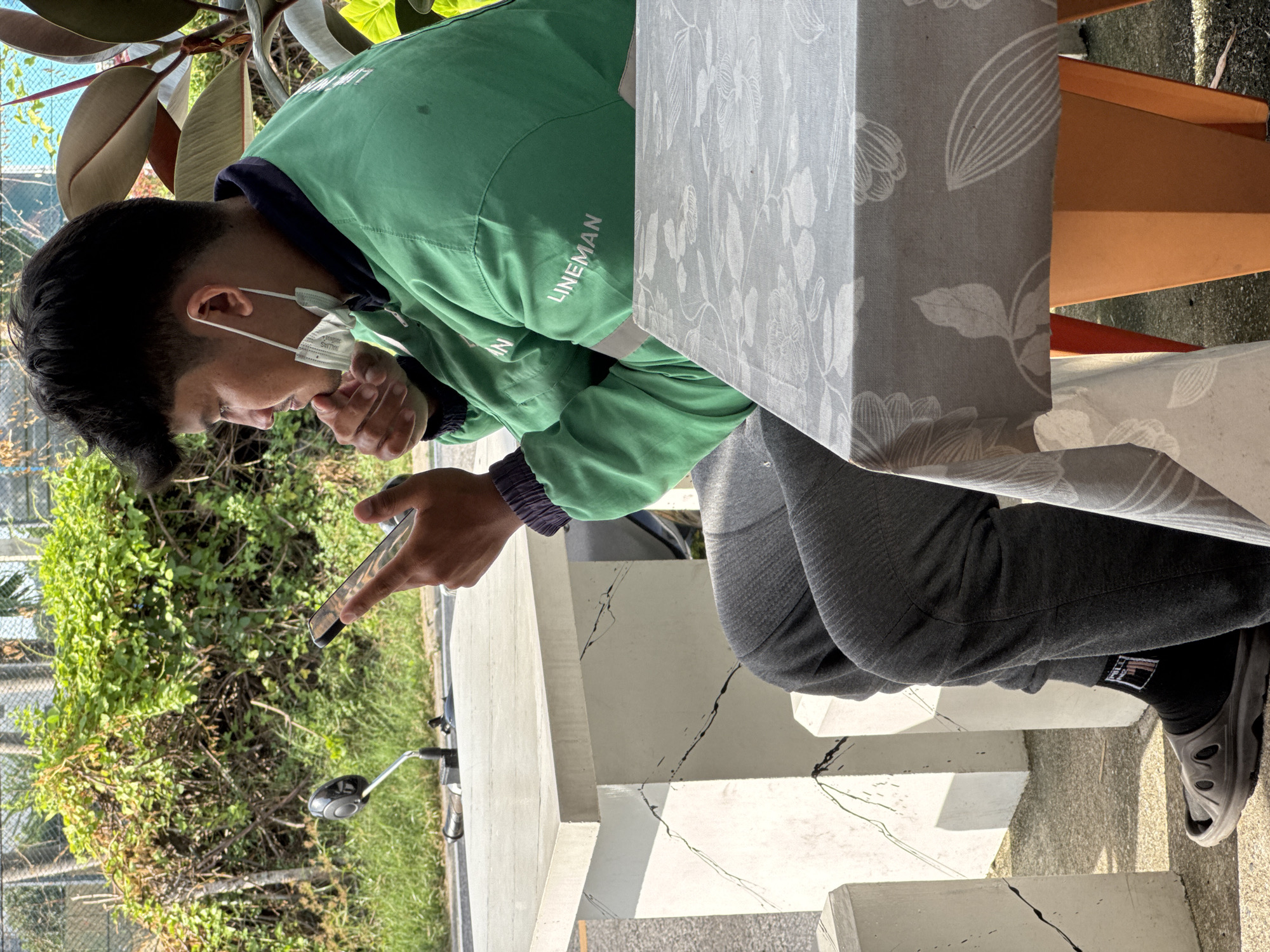 In Phuket City Municipality, Thailand, a young male, appearing to be of Southeast Asian descent, sits at a concrete table with a cracked surface and a second, fabric-covered table in the foreground. He wears a green jacket with "LINEMAN" printed in white on the back, gray sweatpants, black socks with a logo, and dark gray slides. A white surgical mask hangs from his right ear, and he holds a smartphone in his hands, looking intently at the screen. To his left, a motorcycle mirror is visible, suggesting the presence of a motorcycle, and a lush green hedge with some dry branches forms the backdrop. The scene is illuminated by bright sunlight, casting shadows on the ground.