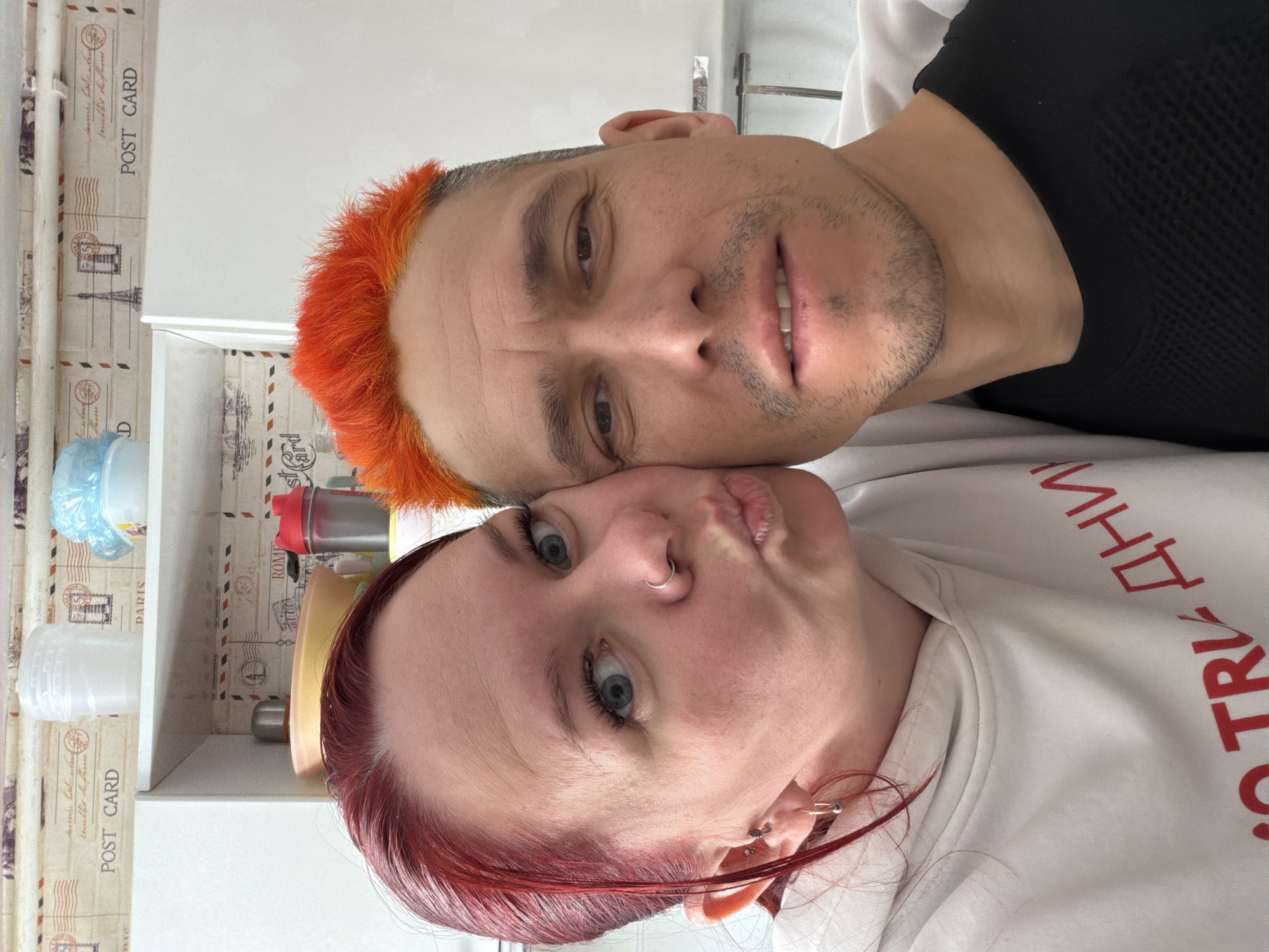 Two individuals, a male and a female, are positioned close to each other, likely in an indoor setting near storage shelves. The male has short, orange-colored hair and is wearing a black top. The female has shoulder length red hair and is wearing a white sweatshirt with Cyrillic text written on it in red. Both are looking at the camera. The location of the photo is presumably in Chaikovsky, Russia.