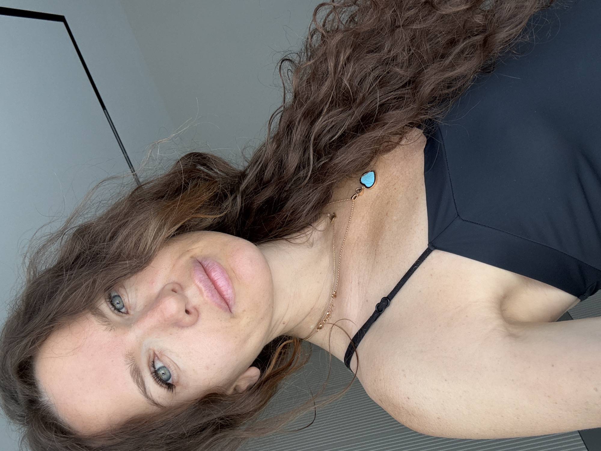 A woman with long, wavy brown hair and blue eyes looks into the camera, her head tilted slightly to the right. She is wearing a black tank top and two delicate gold necklaces, one of which has a small, blue, heart-shaped pendant. Her makeup is subtle, with a touch of pink lipstick. The background appears to be indoors, with a grey, textured wall on the left and a plain grey wall on the right, against which a thin black line, possibly a ceiling beam or part of a light fixture, is visible. There are no identifiable landmarks or clues to indicate the location is Moscow, Russia.