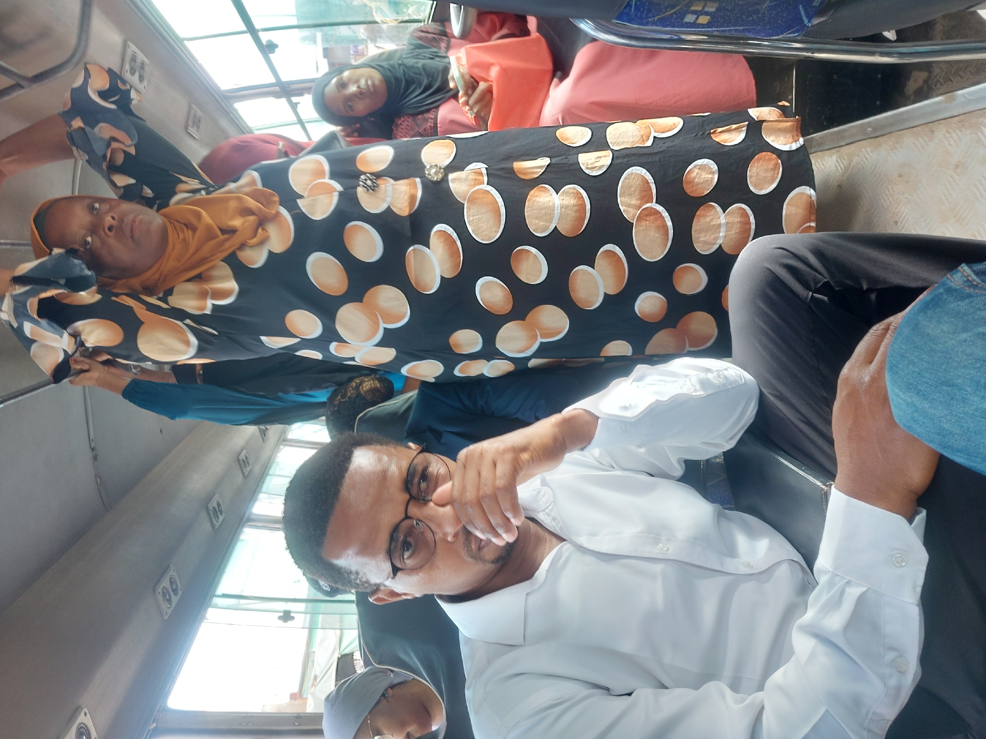 The image shows the interior of a public transportation vehicle in Dar es-Salaam, Tanzania. Multiple people are present.
Foreground: A man wearing a white button-down shirt and glasses is visible.
Middle ground: Another woman wearing a black and brown patterned garment and a yellow head covering is situated behind the man. Further back, another woman is visible in a black head covering.
Background: The interior of the vehicle is seen, showing the window and other passengers.
The lighting appears to be natural daylight.