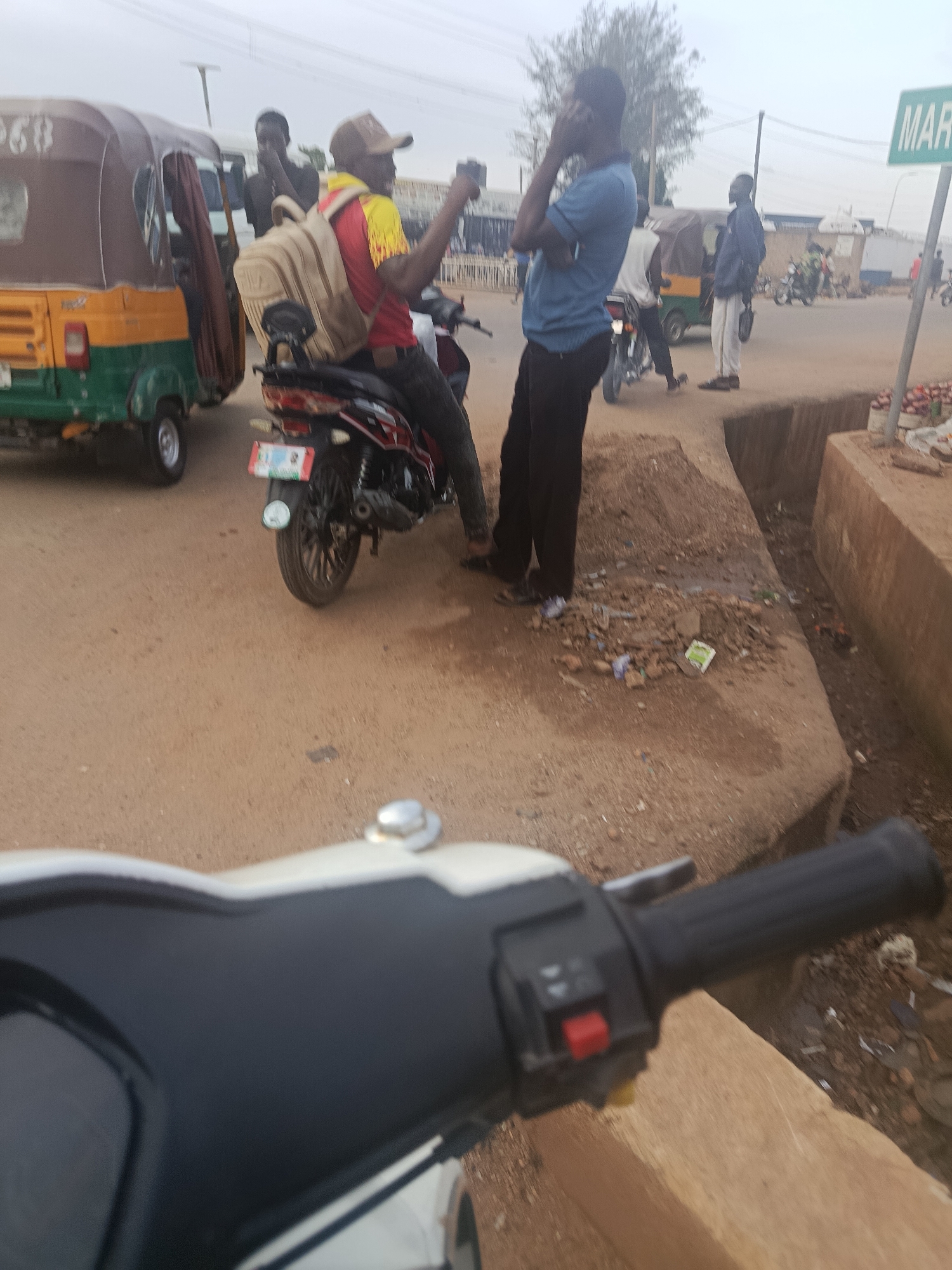 The image depicts a street scene in Jos, Nigeria, likely at a busy intersection or market area. In the foreground, the handlebars and part of the dashboard of a white and black motorcycle are visible, suggesting the photo was taken from the perspective of someone riding or parked on a similar vehicle.
Dominating the mid-ground is a person on a motorcycle, wearing a red and yellow shirt and a beige backpack. This individual appears to be holding up a mobile phone, possibly taking a picture or video. Beside them, a motorized three-wheeled vehicle, commonly known as a "Keke Napep" or "Marwa," is parked. The Keke Napep is predominantly brown and yellow with green accents.
Several other individuals are present in the scene. To the left of the Keke Napep, another person is standing and looking away, partially obscured. To the right of the Keke Napep, two men are visible. One man, wearing a blue shirt and black pants, is holding a phone to his ear, appearing to be on a call. Another man, dressed in a blue jacket and light-colored pants, is standing nearby, holding a dark object, possibly a bag. Further in the background, more people and vehicles are present, including other motorcycles and what appears to be market stalls.
The environment is dusty and appears to be an unpaved or poorly paved road. A visible drainage ditch runs along the side of the road, filled with debris. A green street sign with white lettering, partially visible, reads "MAR," indicating a possible location name or directional marker. The overall atmosphere suggests an active, informal urban setting typical of many Nigerian cities. The sky is overcast, giving the scene a diffused light.