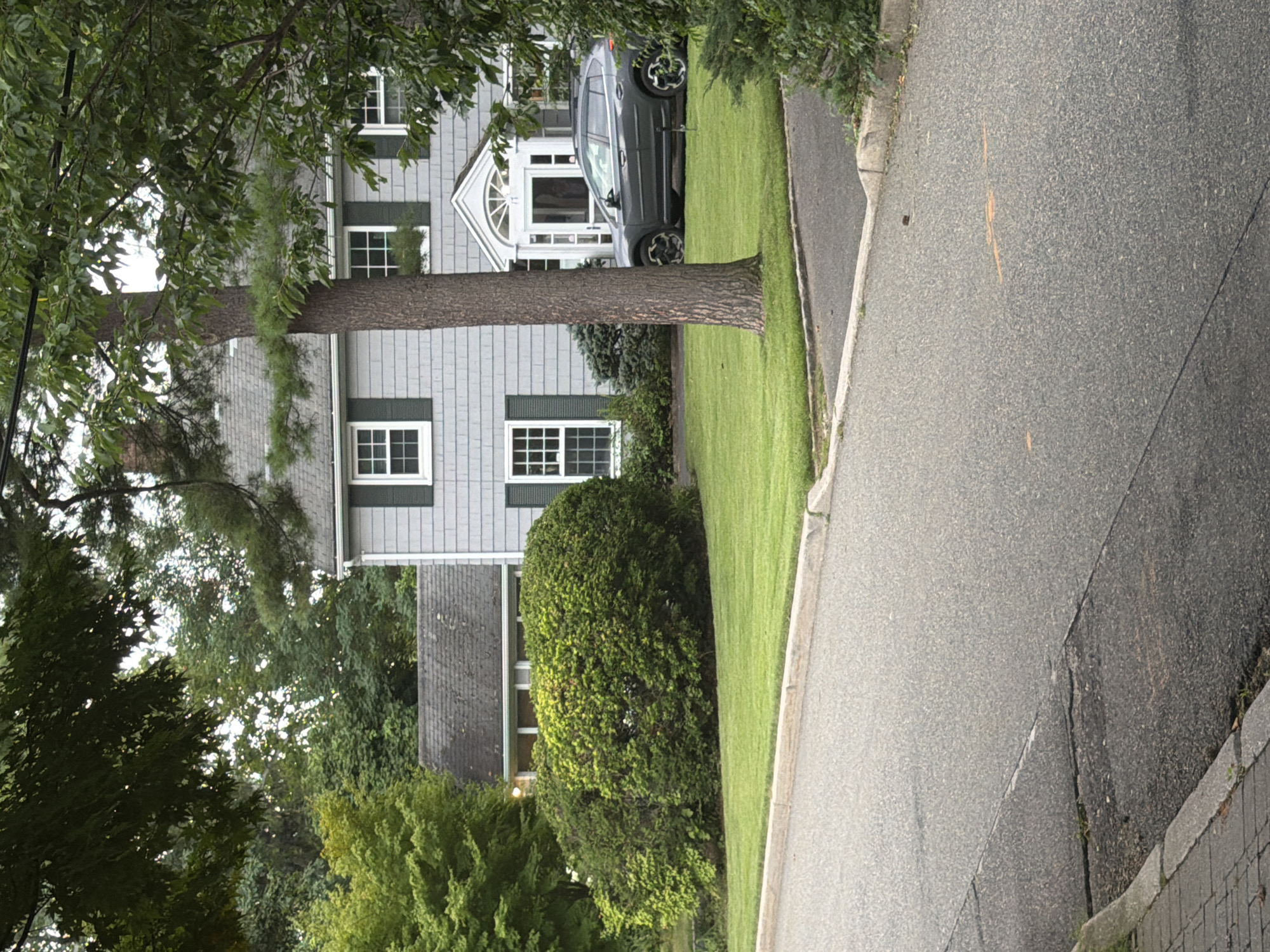 The image shows a two-story gray house with white-trimmed windows and dark green shutters, situated in a suburban neighborhood. A dark gray SUV is parked in the driveway. The house is surrounded by lush green lawn and mature trees, some of which partially obscure the view of the house. The foreground features a paved road with a concrete curb and sidewalk. The overall scene depicts a typical residential setting.