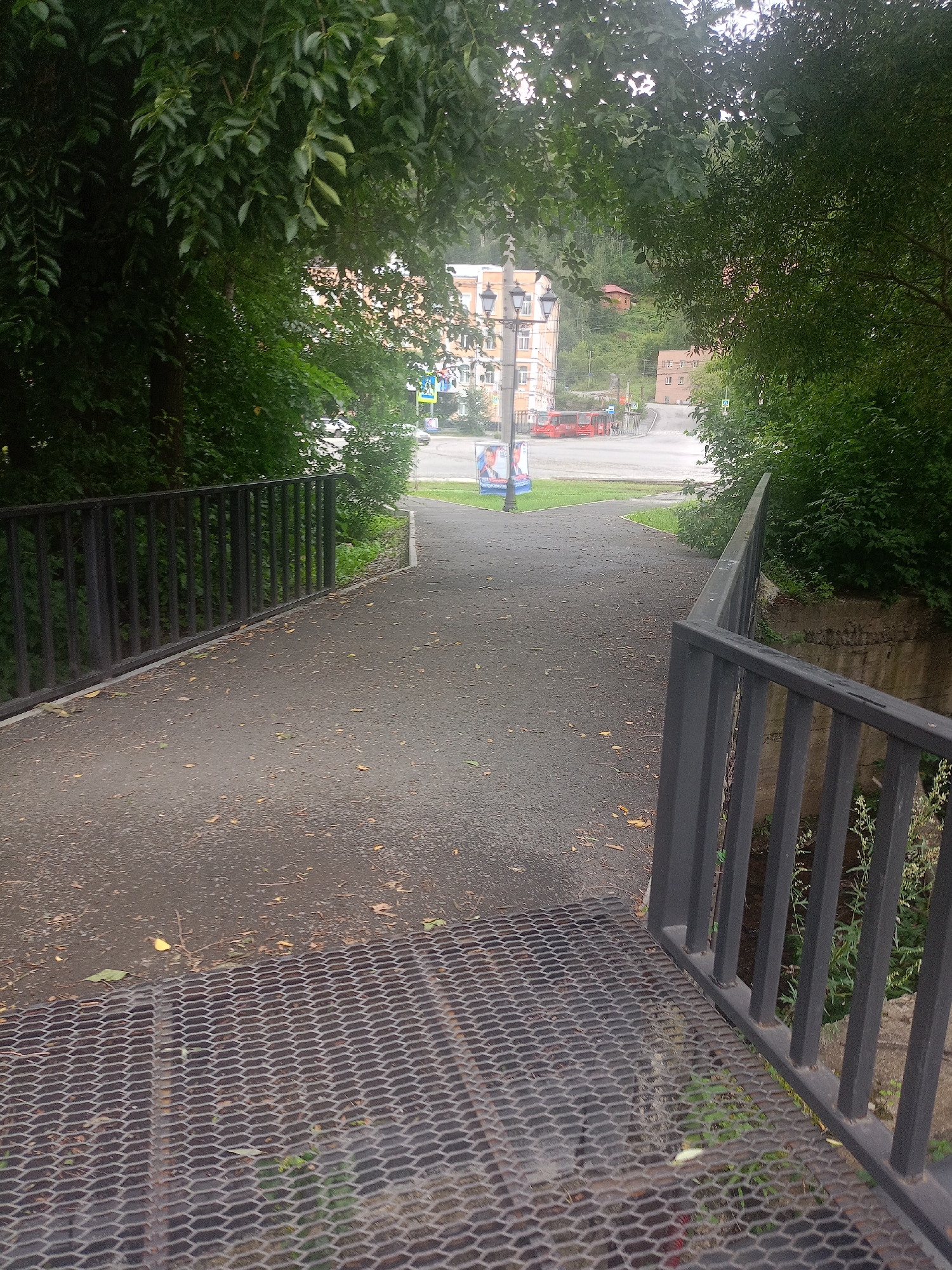 A paved walkway leads towards a street with buildings and a hillside in the background. The walkway is bordered by black metal railings on both sides. In the foreground, a metal grate covers the ground on the right side. On the left, a black metal fence runs along the walkway. In the distance, red buses are visible on the street, and a lamppost with posters stands on the left. The area is surrounded by lush green trees, suggesting a natural setting within an urban environment. The sky appears overcast. This scene is located in Zlatoust, Russia.