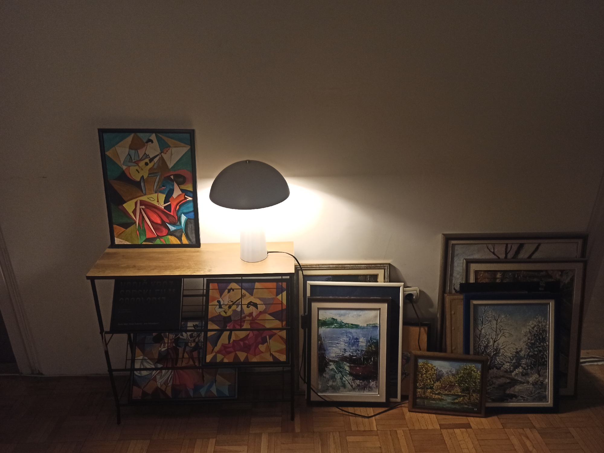 The image displays an indoor scene in Vozdovac Urban Municipality, Serbia, primarily illuminated by a modern desk lamp. In the foreground, a two-tiered wooden and metal table serves as a display surface. On the top left of the table, a framed, colorful cubist painting featuring geometric figures and a guitar stands upright. Adjacent to it, a white-based lamp with a grey, dome-shaped shade casts a bright, warm light upwards and outwards, illuminating the wall and nearby objects.
The lower shelf of the table holds more artworks. On the left, a dark rectangular item displays white text, partially legible as "VOZIC KRAJ" and "SRECKO FILIP 2003 2017". To its right, another cubist-style painting, similar in aesthetic to the one above, is visible.
To the right of the table, a collection of framed landscape paintings is propped against the wall on the wooden parquet floor. These artworks vary in size and style, depicting scenes of nature, including water bodies, trees, and winter landscapes, with some appearing more traditional. The lighting suggests a dimly lit room, with the desk lamp providing the primary illumination, creating a focused and intimate atmosphere for the displayed art. No individuals are present in the scene.