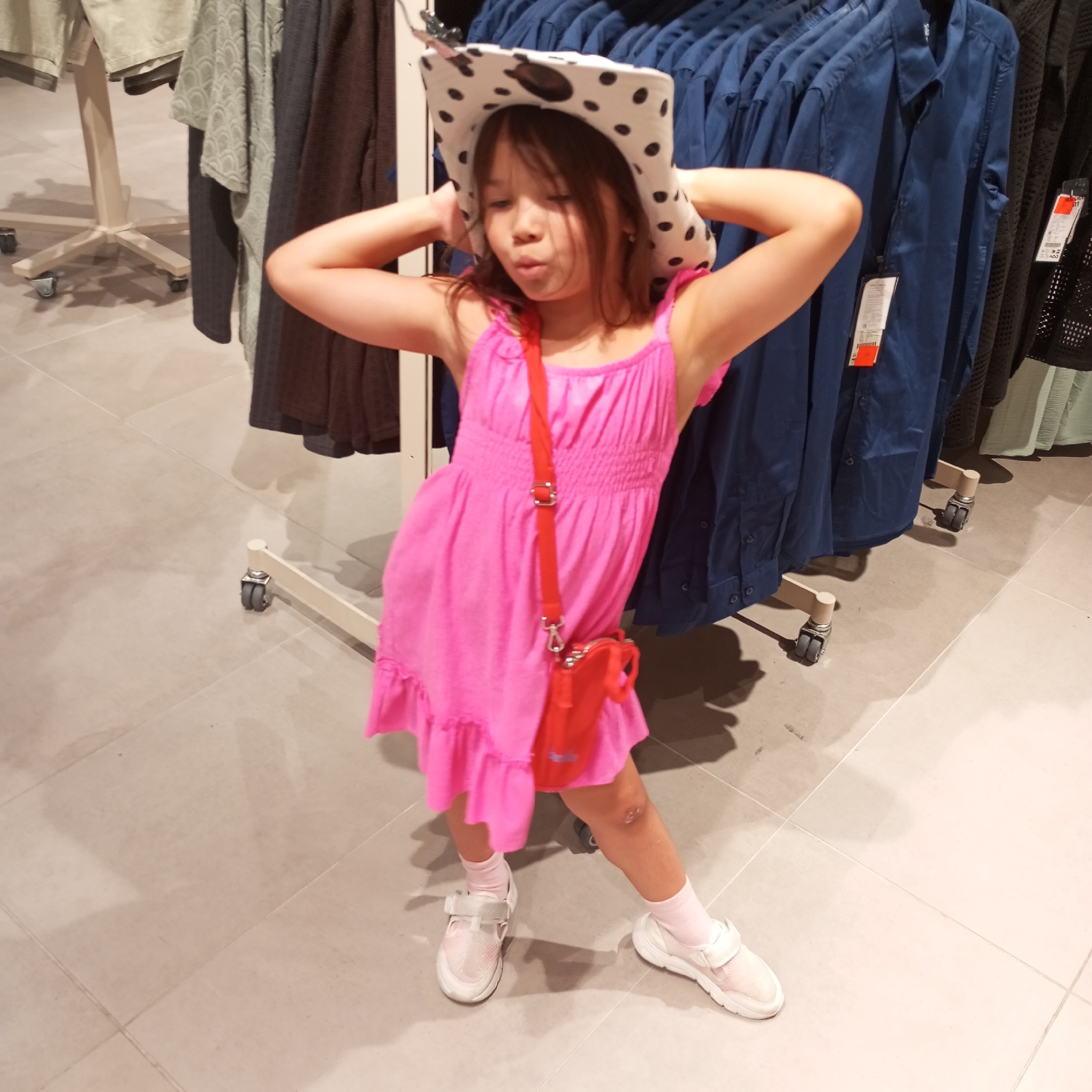 A young girl wearing a pink dress and a spotted hat poses in a clothing store. She is standing in front of racks of clothing, with a red purse slung across her body. Her arms are raised behind her head, and she is looking down with her mouth slightly open. The store appears to be well-lit, with light-colored tiled flooring. The background shows rows of blue shirts on hangers, as well as some other garments on display. The scene suggests a shopping trip, possibly for new clothes. There is no indication of the location being in Sochi, Russia.