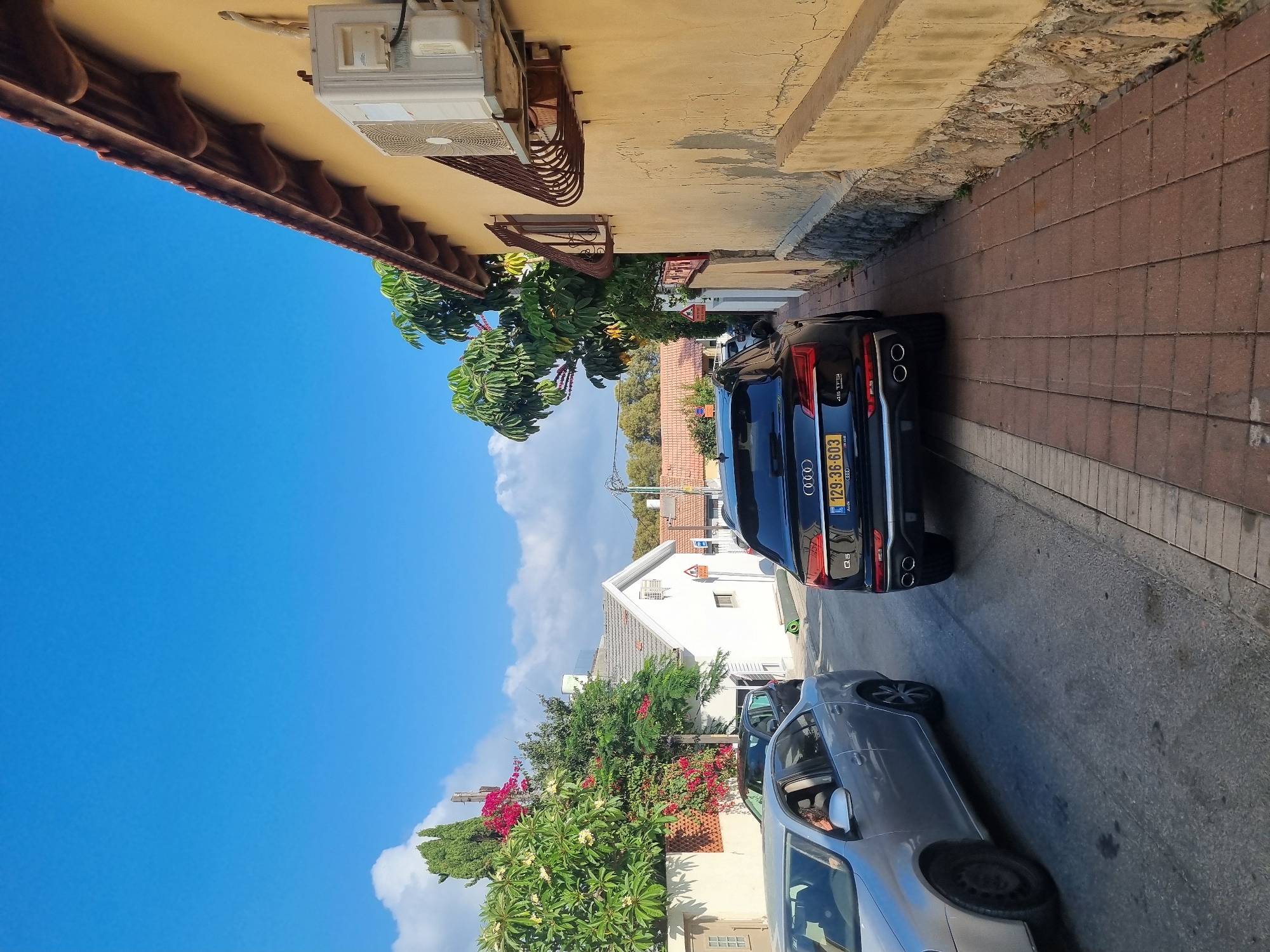 The image depicts a street scene in Ashdod, Israel. In the foreground, a black Audi Q5 is parked on the right side of the road, its rear facing the camera. To the left of the Audi, a silver sedan is also parked. The street is paved, and a sidewalk made of reddish-brown bricks runs along the right side of the road. A yellow building with a textured wall and a brown tiled roof is situated on the right, with an air conditioning unit mounted on its exterior. Trees with green foliage and some with pink and yellow flowers are visible on the left side of the street, behind a fence. White buildings with tiled roofs and a blue sky with scattered clouds are visible in the background. The overall impression is a residential street on a sunny day.