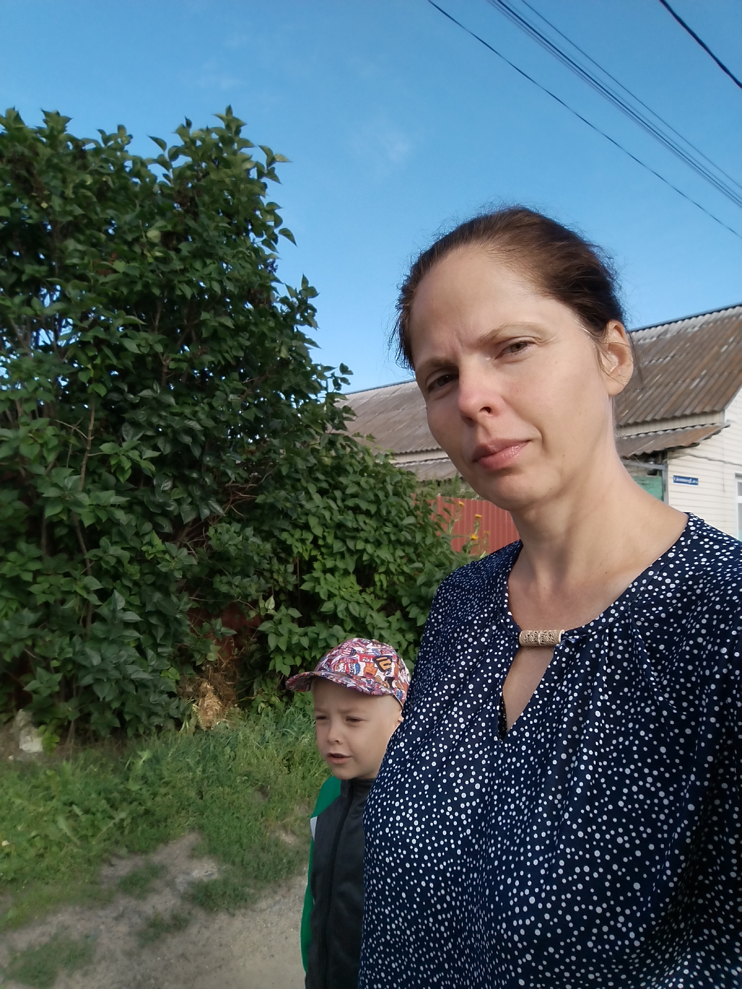 A woman with brown hair pulled back, wearing a dark blue polka-dot top with a decorative clasp, stands in the foreground, looking towards the camera with a neutral expression. To her left and slightly behind her is a young boy wearing a colorful baseball cap and a green and gray jacket. They are outdoors, with a large, leafy green tree to the left of the frame and a house with a corrugated roof in the background on the right. The sky is a clear blue, with visible power lines overhead. The ground appears to be a dirt path with grass and weeds. The image is likely taken on a street or pathway in a residential area, possibly in Zavodoukovsk, Russia, given the architectural style of the house and the general rural/suburban feel of the surroundings.