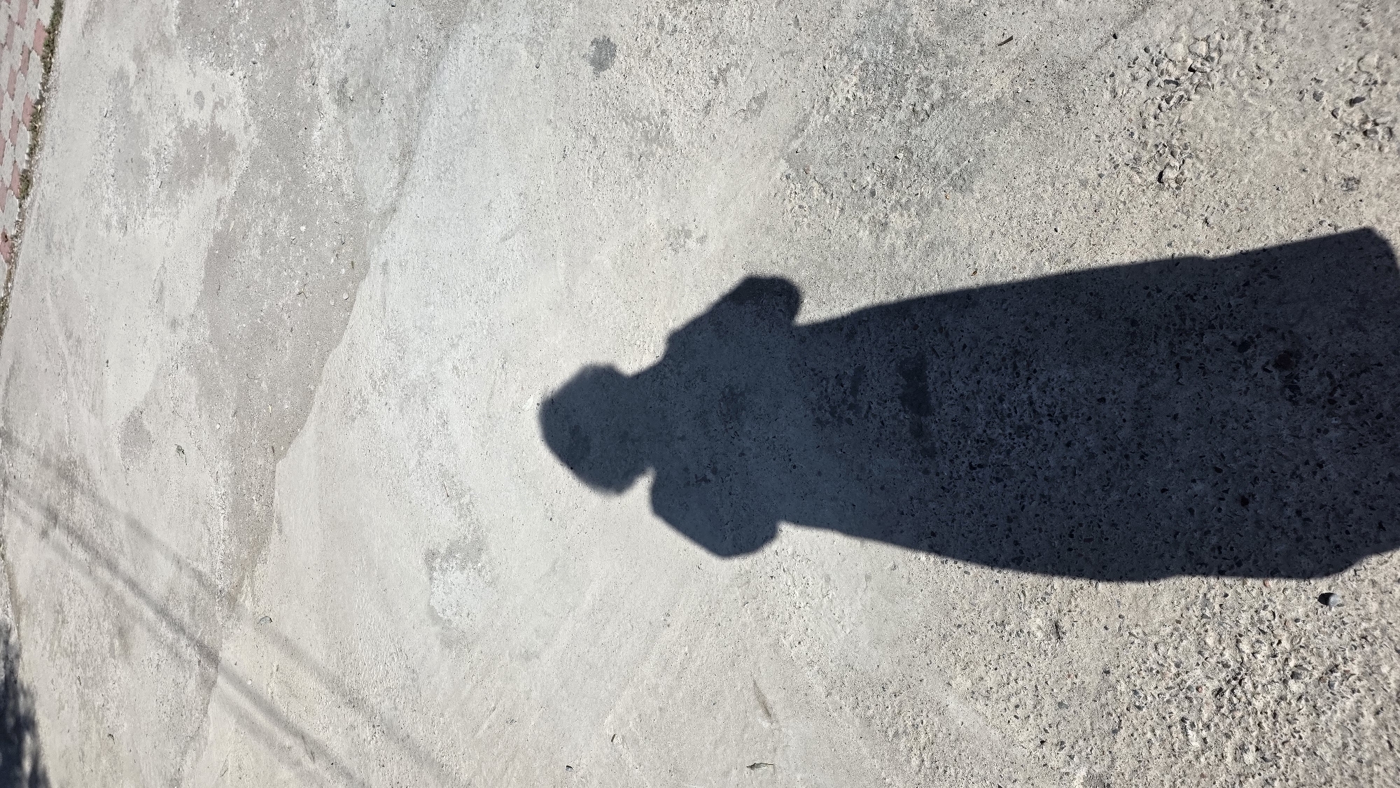 The image shows the shadow of a person standing on a rough, grey concrete surface. The shadow is cast by sunlight from the right, creating a dark silhouette that is elongated and appears to be of a person wearing a long coat and a hat. The concrete surface shows variations in texture and color, with some lighter patches and a scattering of small pebbles and debris. In the upper left corner, there are faint shadows of what appear to be wires or branches from above, possibly indicating the presence of overhead lines or trees. The background consists of more of the same concrete surface, leading to a slightly different texture near the top right edge, where a small patch of red and grey checkered pavement is visible. The overall impression is an outdoor scene, likely in a public space, possibly a sidewalk or courtyard, in Tokmok City, Kyrgyzstan.