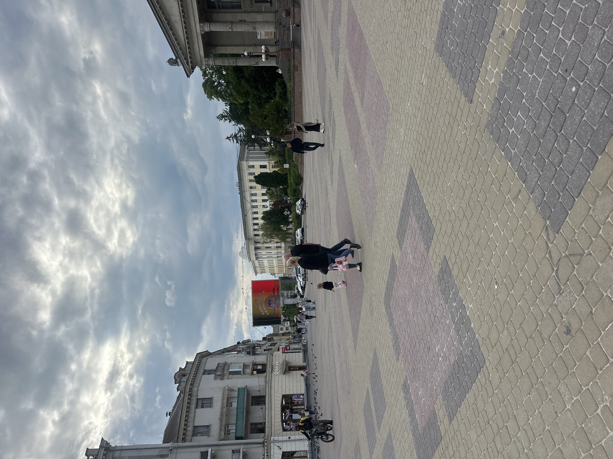 The image depicts a wide, paved plaza in Ternopil, Ukraine, under a cloudy sky. The plaza is laid with interlocking pavers in a geometric pattern of gray, pink, and beige.
In the background, to the left, is a multi-story building with a light-colored facade, featuring several balconies and windows. A sign for "Blisk" is visible on the ground floor. Further to the left, another building with architectural columns is partially visible.
Across the plaza, to the right, stands a large, classical-style building with a prominent colonnade and a grand entrance. Trees and greenery are visible behind this building.
Several people are present in the plaza:
* A person on a motorcycle with a yellow backpack is on the left side of the frame, moving towards the viewer.
* A family, consisting of two adults and a small child, are walking across the plaza, with the adults holding hands. The child is walking slightly ahead.
* Another couple, a man and a woman, are walking together in the middle-right of the frame.
* Two other individuals are walking in the middle-left of the frame.
* A group of people are gathered in the distance near the classical building.
A large digital billboard with red and yellow content is prominently displayed in the mid-ground, between the buildings. Pigeons are scattered throughout the plaza. Cars are visible parked on a street in the background. The overall impression is of an urban public space on a potentially overcast day.