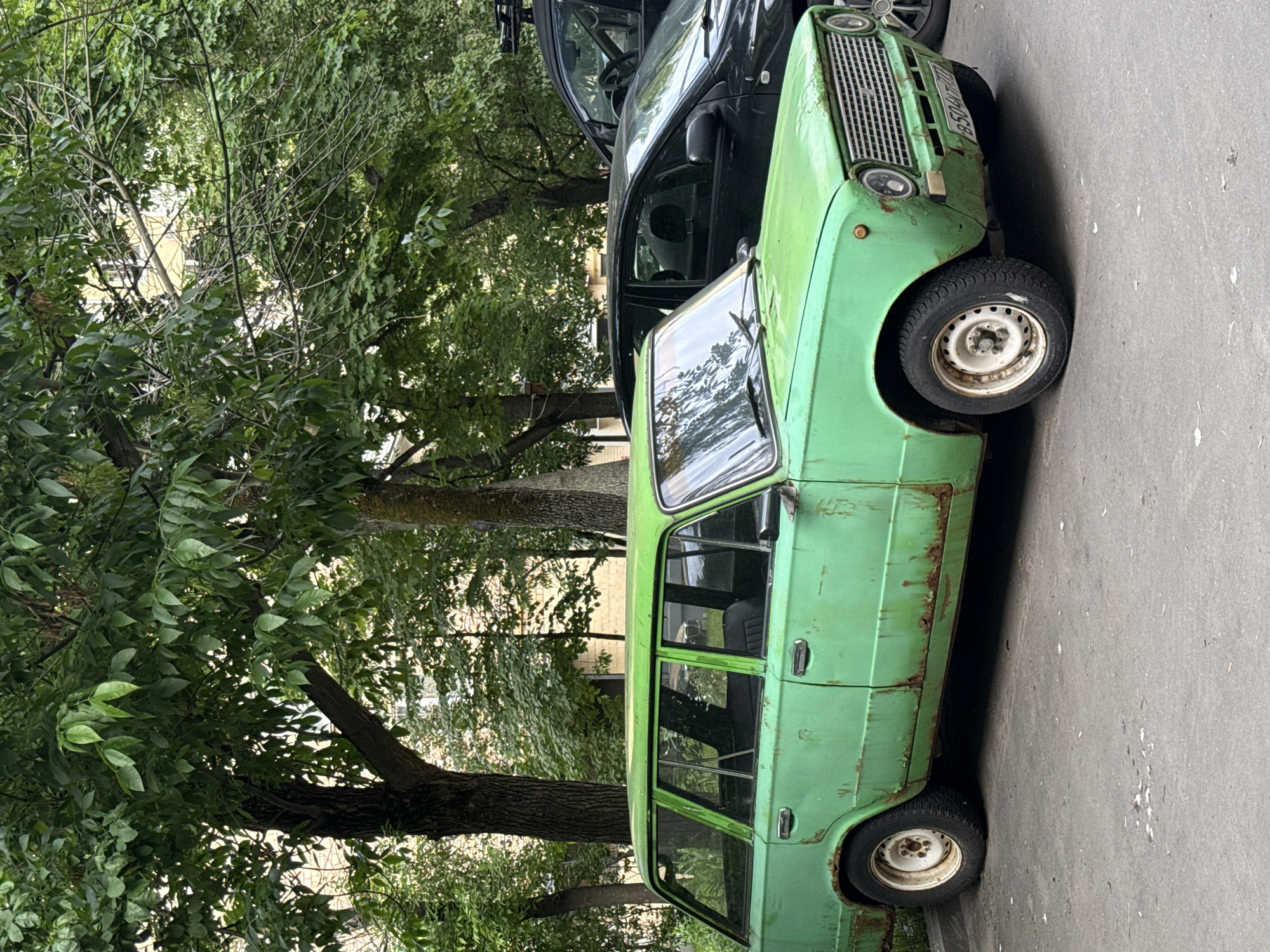 This outdoor daytime photograph, rotated 90 degrees, captures a street scene featuring two parked cars next to a densely treed area. The primary subject is a weathered, light green vintage station wagon, identifiable as a Lada (likely a VAZ-2102 or similar Zhiguli model), parked on grey asphalt pavement. The car shows extensive signs of age, including significant rust along its body panels, especially above the wheel wells, and faded, peeling paint. Its front grille, circular headlights, and a front license plate reading "B504Y0" are visible, though partially obscured and worn. The car has white steel wheels.
Behind and to the left of the green Lada, a darker, more modern sedan (possibly black or dark blue) is partially visible, showing its front-left section and windshield. The left side of the image is filled with lush green foliage from several trees, with visible tree trunks suggesting a treelined street or adjacent park. Faint, light-colored buildings can be discerned through the leaves in the background.
No people are present, and no specific activity is taking place; the cars are simply parked. The lighting indicates it is daytime, and the weather appears clear. The style of the older car and the urban environment are consistent with the Moscow, Russia location context. The scene conveys a sense of quiet urban life, highlighted by the enduring presence of a well-used vintage vehicle.