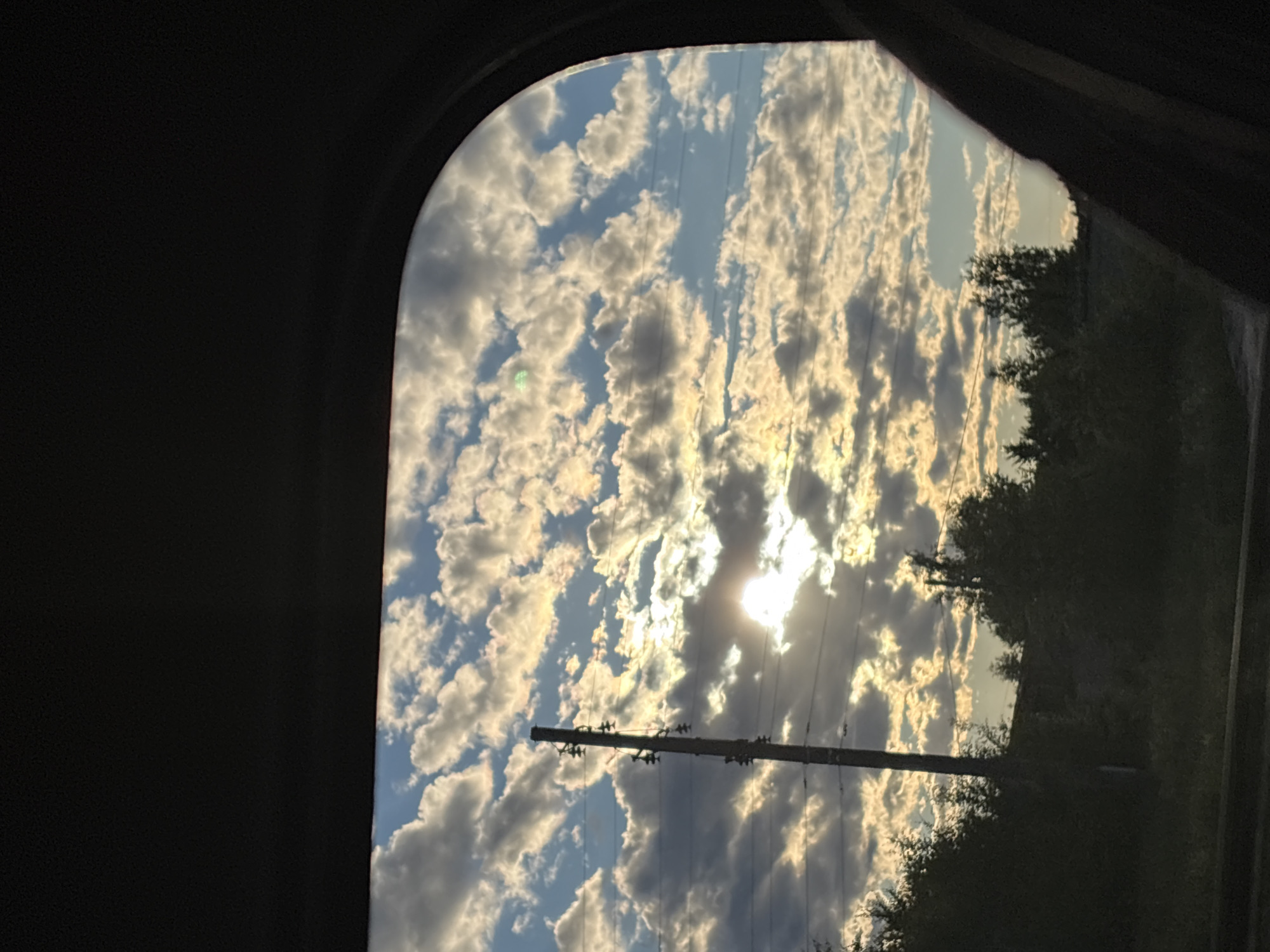 The image shows a view of the sky and trees through a vehicle window. The sky is filled with cumulus clouds illuminated by the setting sun, which is partially obscured by the clouds. A telephone pole with insulators and wires stands vertically in the left portion of the frame. The foreground is dark, suggesting the interior of a vehicle. Trees and foliage are visible at the bottom of the frame, partially silhouetted against the sky. The scene appears to be a rural or suburban landscape, potentially on the outskirts of Makaryev, Russia, given the presence of the telephone pole and the natural surroundings.