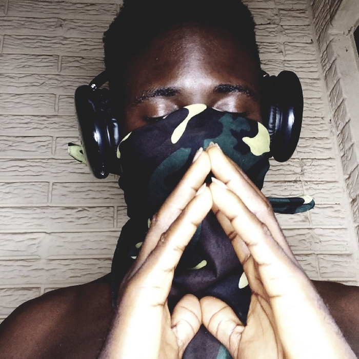 A forensic description of an indoor scene in Narayi, Nigeria. The image features a single, dark-skinned adult male with short, dark hair, positioned against a light-colored brick wall. His eyes are closed. He wears black over-ear headphones and a camouflage-patterned fabric covers his lower face, obscuring the nose and mouth. Both hands are raised in front of his face, fingers interlocked and pressed together to form a prominent triangular shape. His bare shoulders are visible. The brick wall behind him exhibits a light-colored stretcher bond pattern.