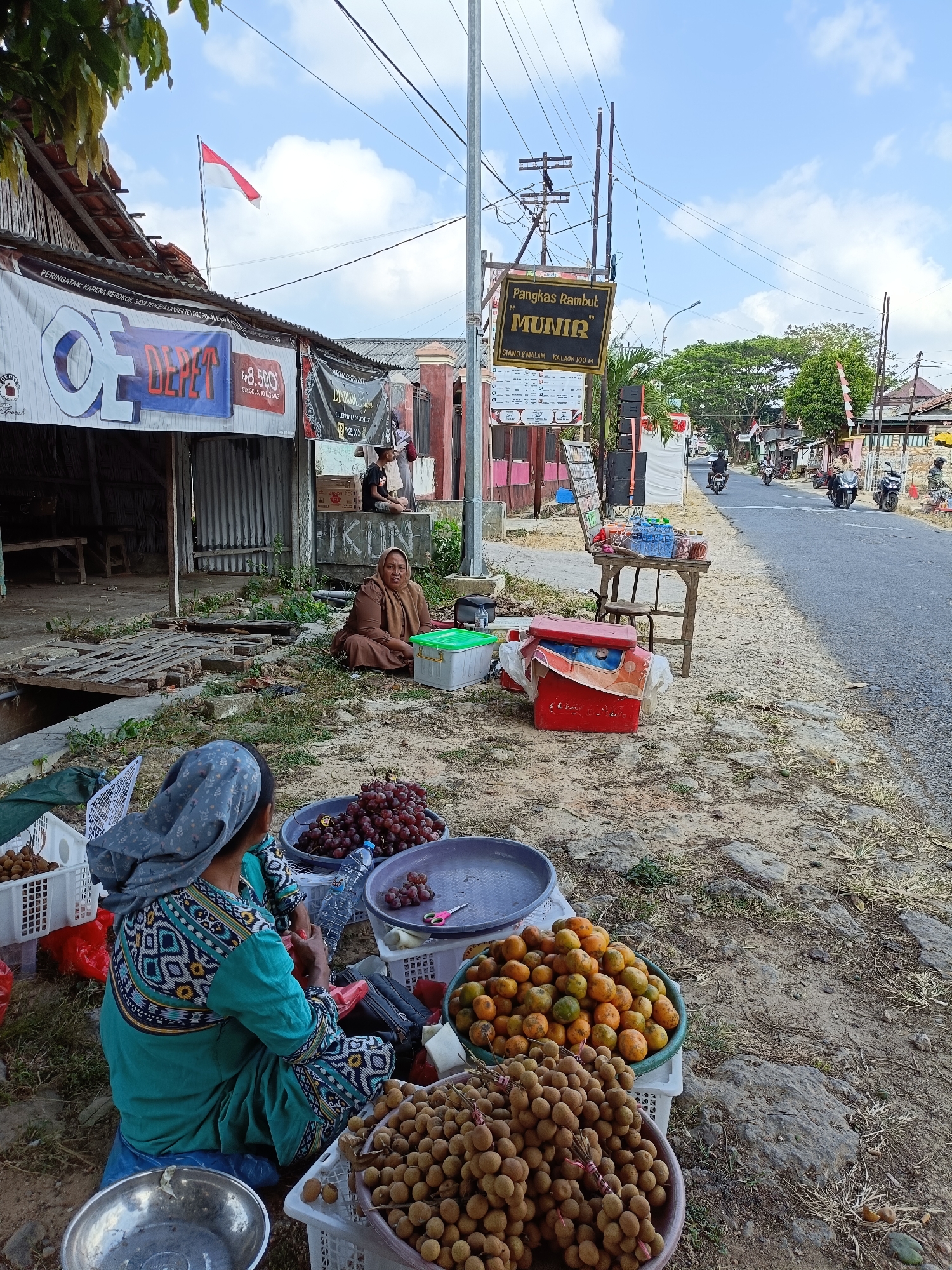 A street scene in Pordapor, Indonesia, depicts a roadside fruit stand. Two women are present, one sitting and facing away from the camera, wearing a patterned headscarf and a teal and patterned outfit, and another sitting to her right, facing the camera, dressed in a brown headscarf and brown clothing. In front of the seated women are baskets of fruit, including grapes, longans, and oranges. A small table to the right holds bottles of water and other items. Behind the fruit stand, a street is visible with motorbikes and pedestrians. Overhead, power lines crisscross the sky, and a red and white Indonesian flag flies from a building. The area appears to be a commercial street with various shops and stalls.