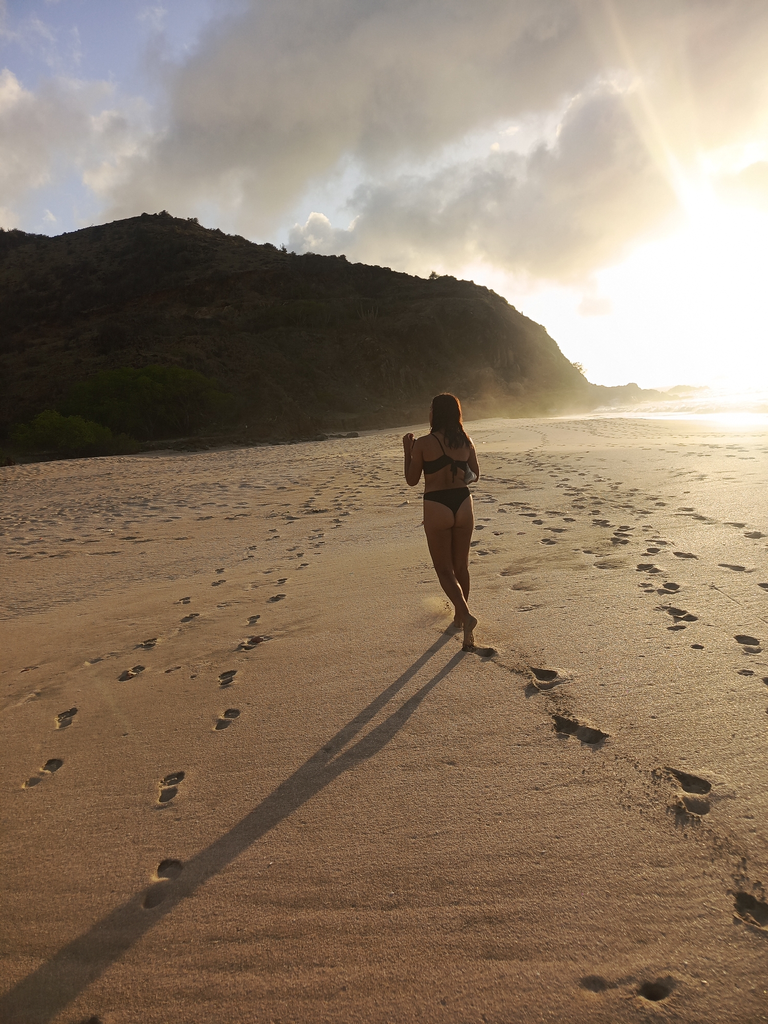 A woman in a black bikini walks away from the camera on a sandy beach during sunset in Pedro González, Venezuela. The sun is setting behind a hill on the right side of the image, casting a warm glow over the scene and creating long shadows on the sand. The beach is covered in footprints. The ocean is visible in the distance, with waves crashing on the shore. The sky is cloudy, with the sunbeams breaking through the clouds.