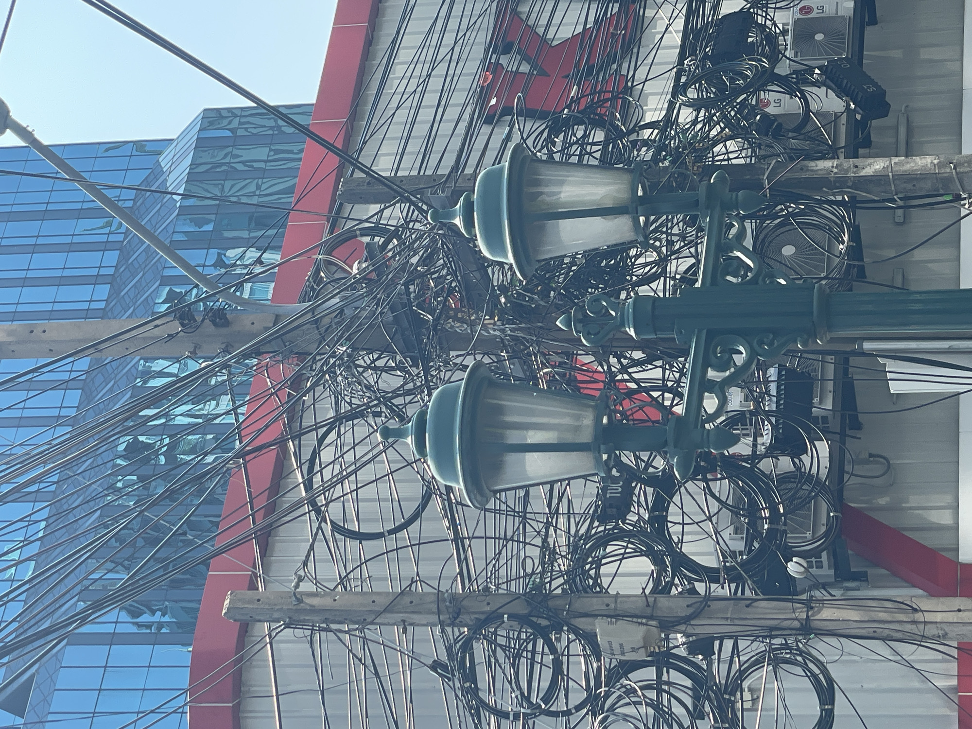 This image captures a dense and chaotic network of overhead utility wires and communication cables in an urban environment, indicative of Bangkok, Thailand. The multitude of black wires are intricately tangled and crisscross across the frame, supported by wooden utility poles and crossarms.
Prominently featured are two ornate, dark green streetlights or lampposts with classic designs, their translucent shades reflecting the bright light. These lights are positioned amidst the thick cluster of cables.
In the background, a modern building with a reflective blue glass facade is visible on the left, mirroring the clear blue sky. This contemporary architecture provides a stark contrast to the older, more complex utility infrastructure. To the right, behind the web of wires, is another building with a red and white/grey facade. Several outdoor air conditioning units, some marked with the "LG" logo, are mounted on this structure.
The scene is bathed in bright daylight under a clear sky. While no human activity is depicted, the image highlights the bustling and complex infrastructure of a modern city. Visible text includes "LG" on air conditioning units and partial text like "COAXIAL" and "TUC" on some cables or equipment. The overall impression is one of dense urban development where utility lines form an overwhelming visual element.