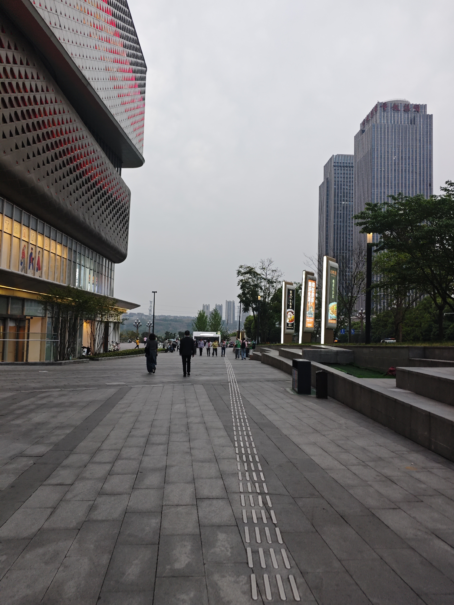 The image shows a street scene in the Liangjiang New Area, Chongqing, China. A paved walkway with tactile paving for the visually impaired stretches into the distance. On the left is a large, modern building with a distinctive geometric facade featuring triangle-shaped perforations and illuminated storefront windows. Several people are walking along the walkway, including two figures in the foreground. On the right, there are several illuminated advertising signs and landscaped areas with trees. In the background, several tall buildings can be seen under an overcast sky. The general scene depicts a modern urban environment.