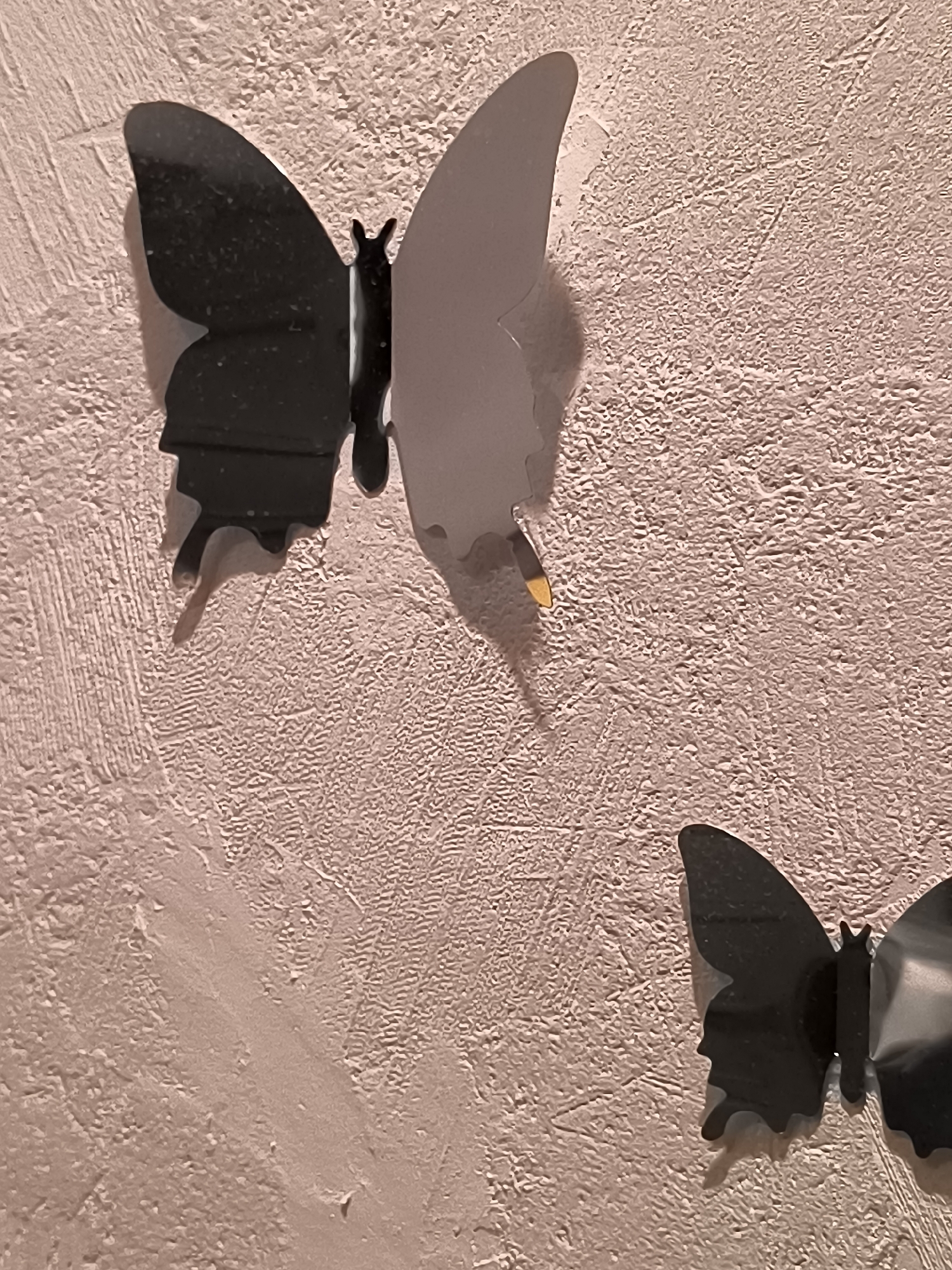 The image displays two black butterfly decorations attached to a textured, pale pink wall. The butterfly on the left is larger and is split in half, with the left wing being black and the right wing appearing to be a lighter, perhaps grey or white, color. A small gold accent is visible on the lighter wing. The butterfly on the right is smaller, entirely black, and is positioned lower and to the right of the larger butterfly. The wall has a rough, bumpy texture. There are no people or animals present in the image. The location of this scene is not discernible from the image itself.
