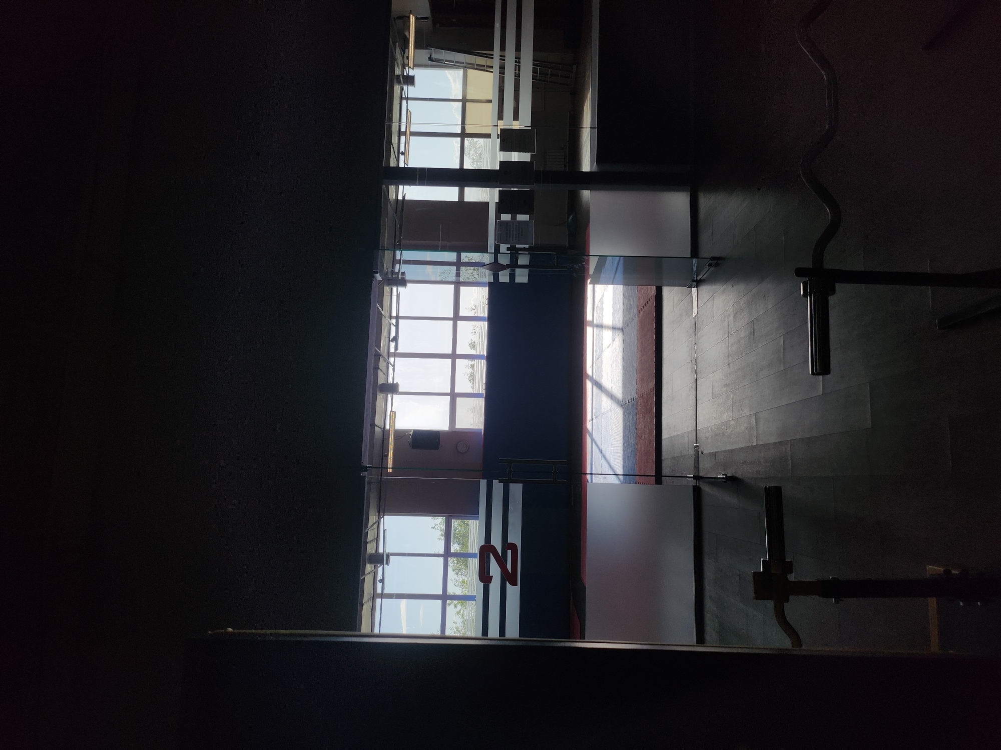 A dim interior view of a building in Unknown, Unknown. The space features a wall with a series of windows that reveal an outdoor view, potentially of a city landscape or park. A prominent "N" logo is visible on a glass partition. The floor appears to be tiled, and the light entering from the windows casts shadows across the surfaces. Workout equipment, including a barbell bar, is visible on the right side. The room seems to be a gym or fitness center.