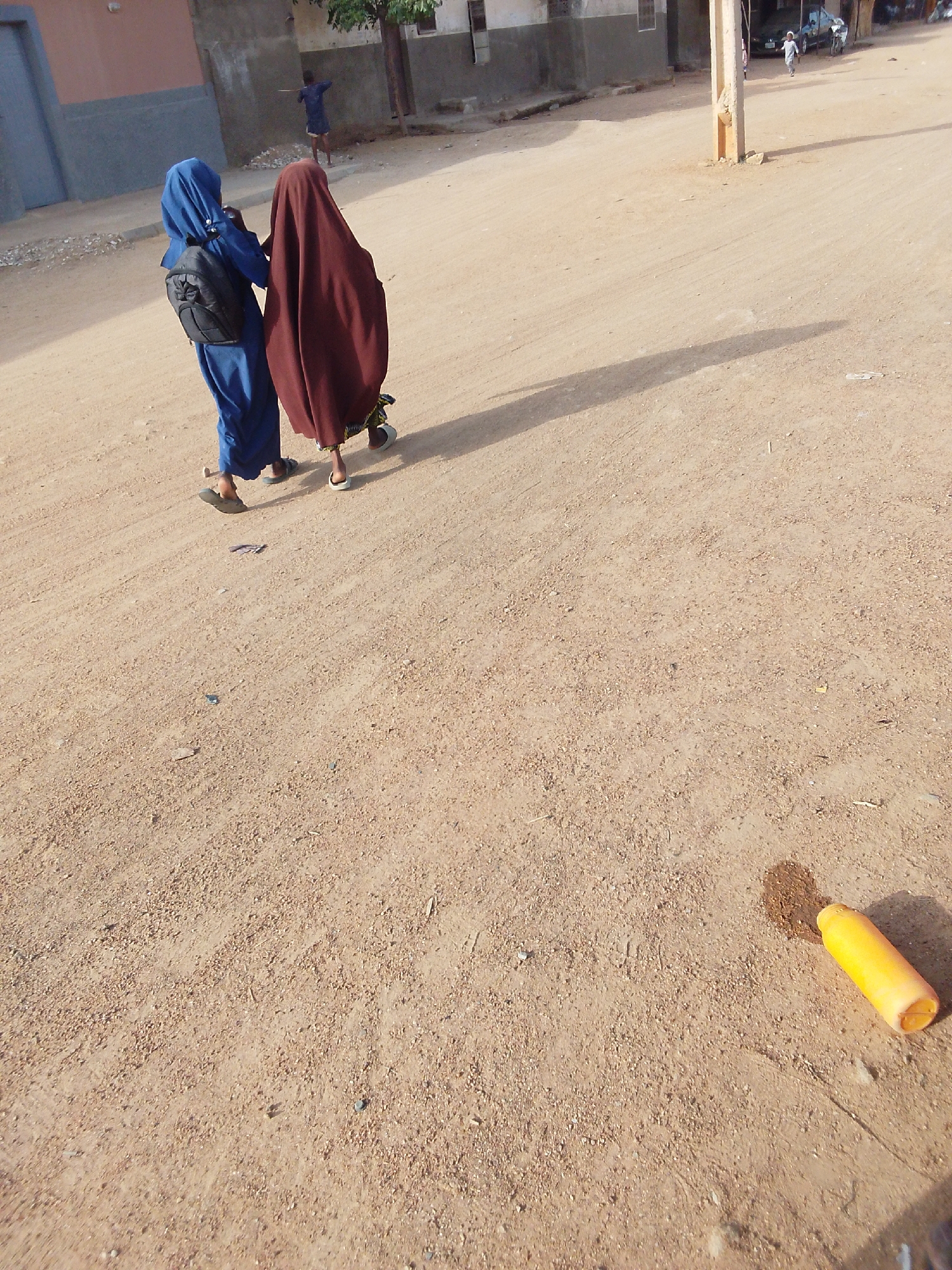 Two females wearing religious attire walk down a dirt street in Bauchi, Nigeria. One is wearing a blue robe and the other a brown robe. A child stands in the background near a building. A yellow bottle lies on the ground in the lower right corner. A utility pole casts a shadow.