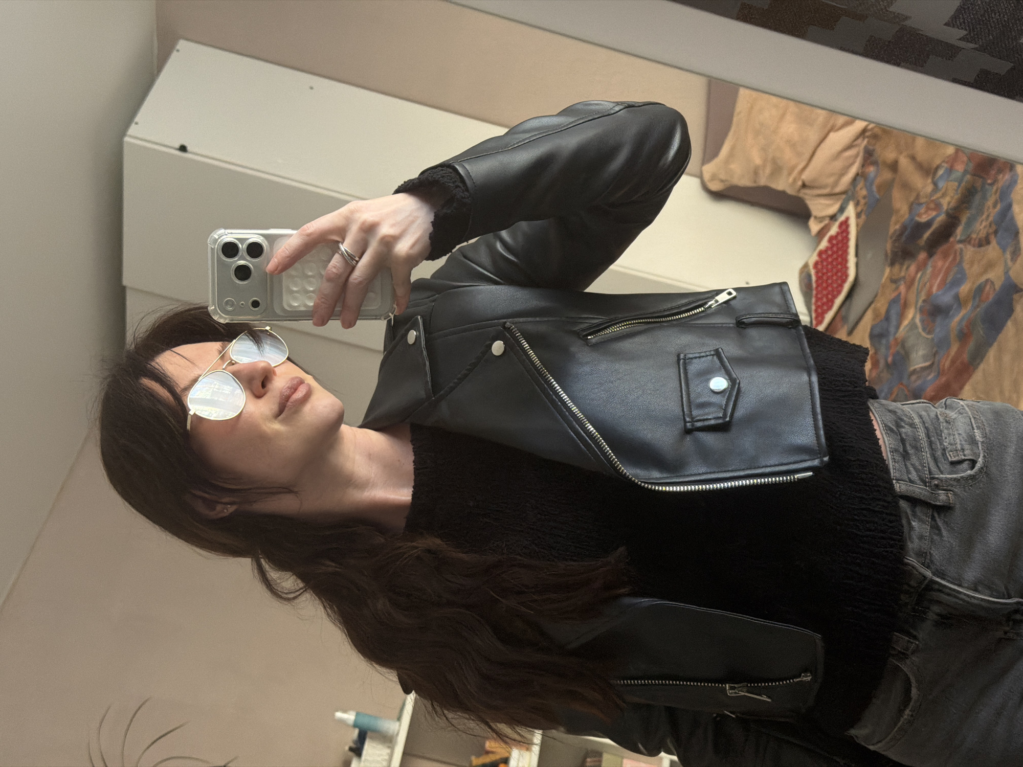 A woman is taking a selfie in a room with neutral-toned walls. She is wearing a black leather jacket over a black, textured top, and grey jeans. She has long, dark wavy hair and is wearing round, wire-frame glasses. She is holding a smartphone with a clear case. The background includes a mirror, reflecting a patterned surface, and a shelf with various items. There is no information to confirm the location of Perm, Russia.