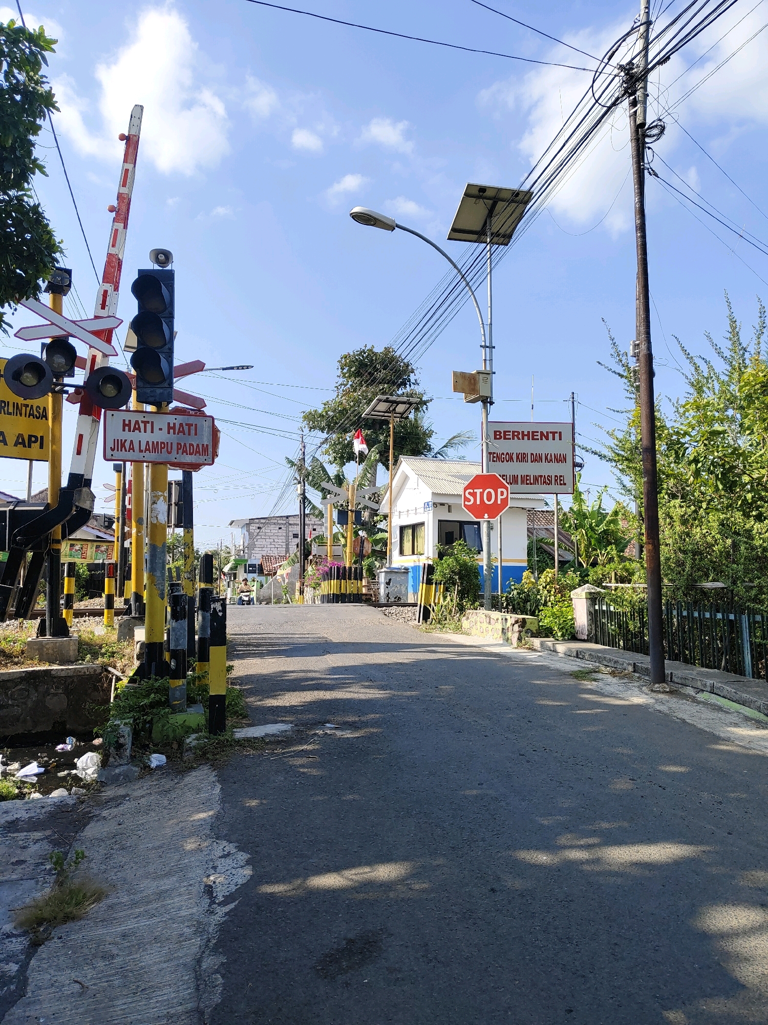 The image depicts a railway crossing on a road in Probolinggo, Indonesia. The scene is characterized by a clear blue sky with scattered clouds and lush green vegetation lining the road. Several traffic signals and warning signs are visible. A railway barrier, painted red and white, is raised, indicating that trains are not currently passing. Traffic lights, displaying red, yellow, and green signals, are mounted on a pole, along with a sign that reads "HATI-HATI JIKA LAMPU PADAM" (Be careful if the lights are out).
Further down the road, another set of signs advises drivers to "BERHENTI, TENGOK KIRI DAN KANAN SEBELUM MELINTASI REL" (Stop, look left and right before crossing the tracks), accompanied by a red octagonal "STOP" sign. A small white building, possibly a signal box or a small station, is situated at the crossing.
The road itself is paved and appears to be well-traveled. The foreground shows a concrete drainage ditch with some litter, including plastic bags and paper. The overall environment suggests a typical Indonesian town or village setting. A motorcycle is visible in the distance, heading towards the crossing. Power lines are strung across the sky, supported by wooden and metal poles. The presence of solar panels on one of the poles suggests a modern infrastructure element.