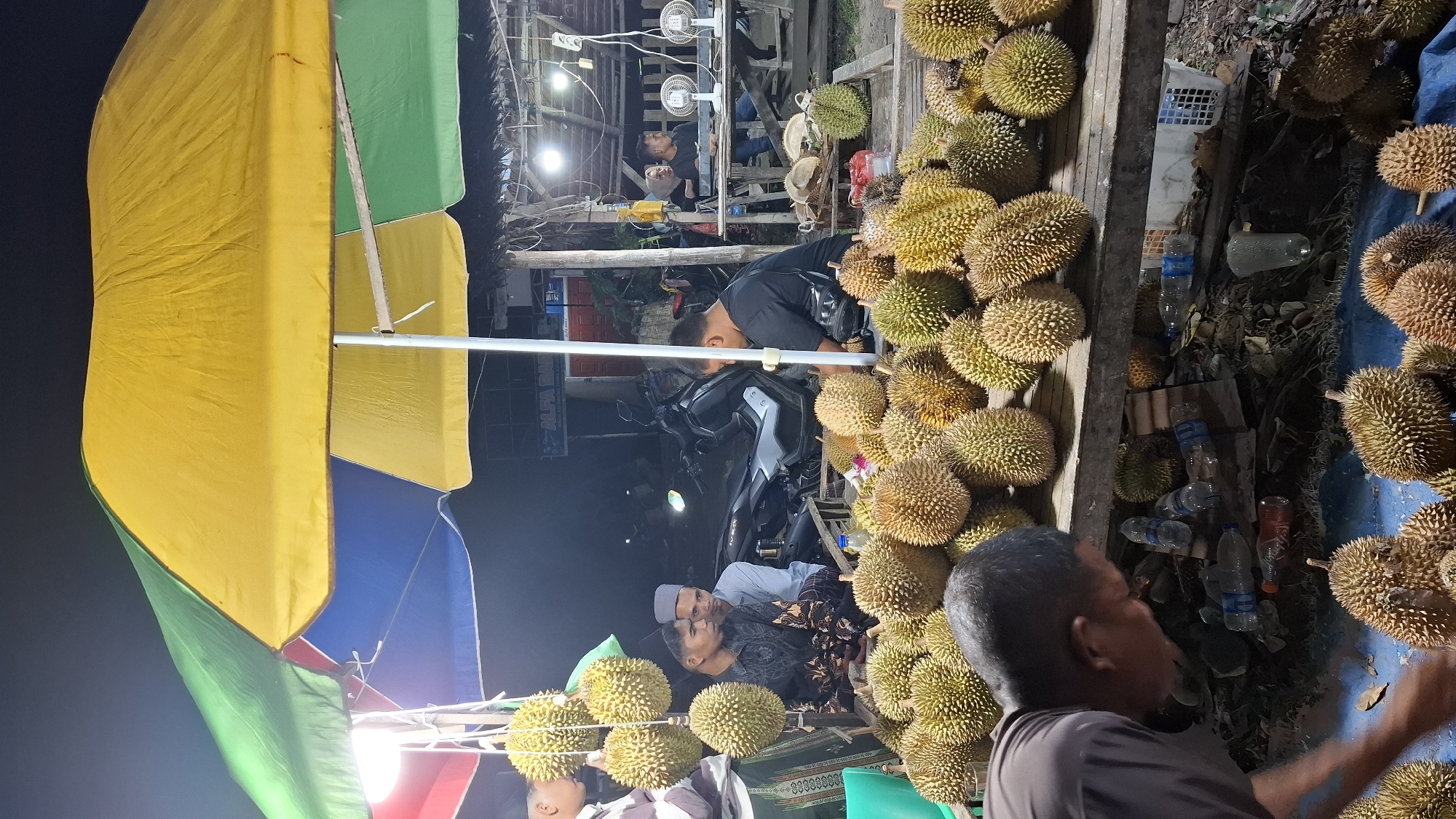A night market stall in Peurupok, Indonesia, is laden with durian fruit under a large, colorful umbrella. Several people are visible in the scene. In the foreground, a man in a grey shirt is bent over, appearing to interact with durian fruit on a blue tarp. Behind him, a table is piled high with durian, with more fruit hanging from above. In the middle ground, three men are seated, two of whom are wearing traditional Islamic skullcaps. A motorcycle is parked behind them. Further back, other people are seated at tables, some with electric fans. The stall is lit by artificial lights, creating a warm and inviting atmosphere despite the darkness of the night. The overall impression is of a lively and bustling local market.