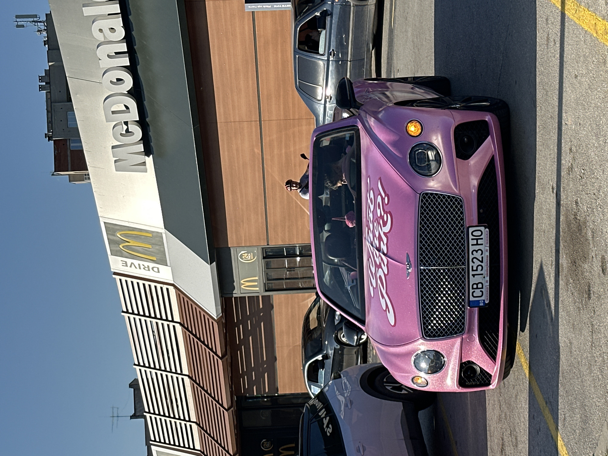 A pink Bentley convertible is parked in front of a McDonald's restaurant in Veliko Tarnovo, Bulgaria. The car has "What the Pink?!" written in large white script on the hood. The license plate reads "CB 1523 HO". Two people are visible inside the car, a driver and a passenger. In the background, there are other cars, including a black BMW and a dark blue Volkswagen, and a person on a scooter. The McDonald's building has its iconic golden arches and "McDonald's" lettering visible, along with "DRIVE" signage. The sky is clear and blue, and the sun is shining.