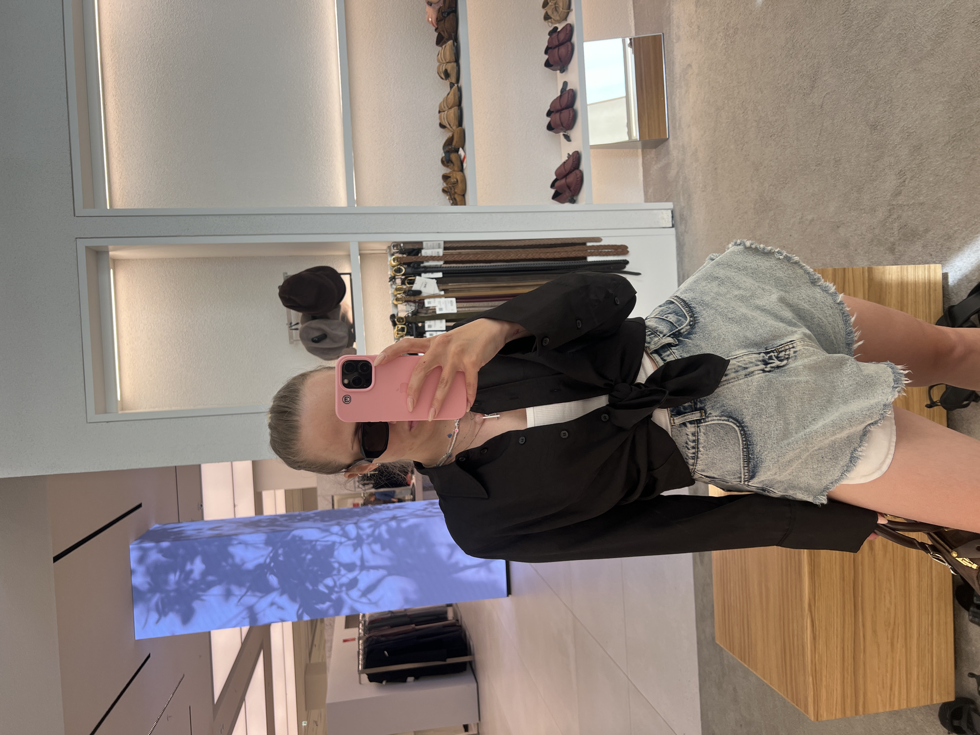 The image shows a young woman taking a mirror selfie in a retail store. She is wearing a black unbuttoned shirt tied at the waist, a white tank top, and light-wash denim shorts with frayed edges. She is also wearing sunglasses and several necklaces. She is holding a pink smartphone. In the background, there are clothing racks, shelves displaying shoes and belts, and a large illuminated pillar with a projected image of leaves. The floor is carpeted in some areas and tiled in others. A wooden display table is visible to the left of the woman. The location is identified as a retail store in Saint Petersburg, Russia.