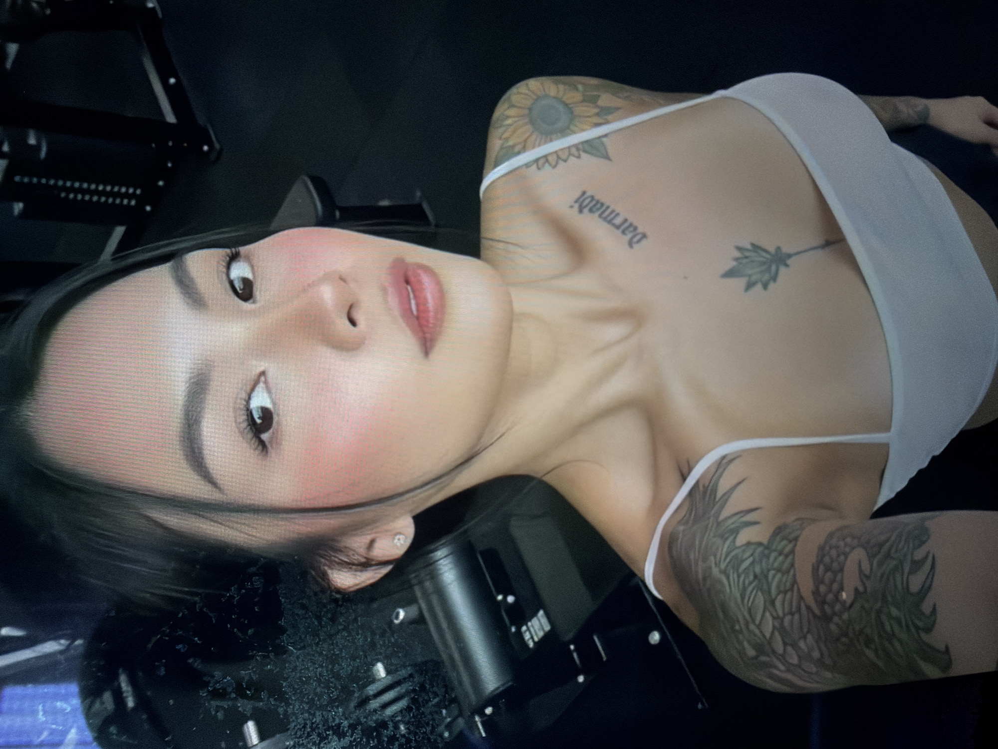The image shows a close-up of a woman with dark hair pulled back, wearing a white tank top. She has visible tattoos on her chest and arms, including a sunflower on her right shoulder and a dragon on her left arm. She is wearing light makeup, with blush on her cheeks and lip gloss. The background is dark and appears to be a gym, with equipment visible. The image is taken from a selfie perspective. There is no information in the image to suggest a location in the city of Shumen, Bulgaria.