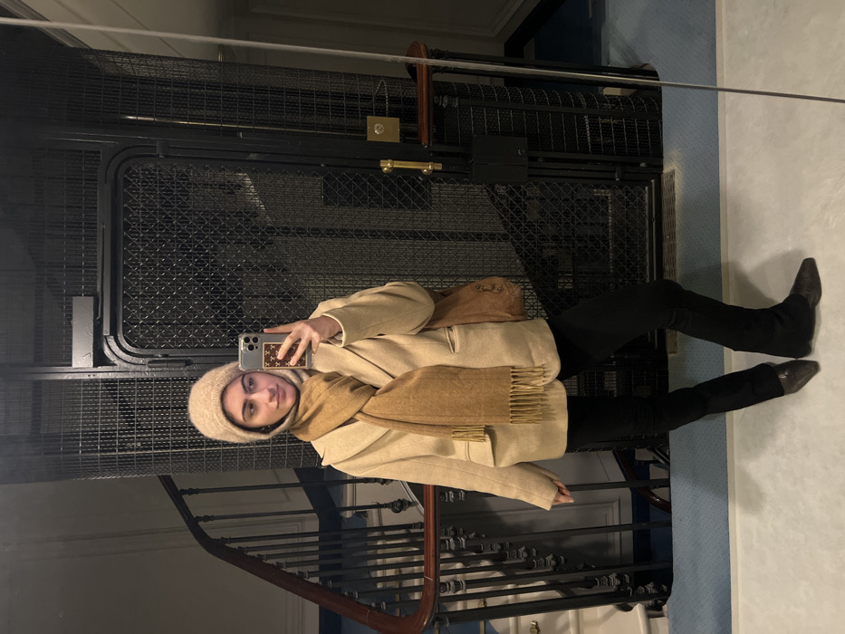A person takes a mirror selfie in an old elevator or stairway with a wrought iron gate. They wear a cream-colored coat with a brown scarf and hat. Black pants and dark boots complete the outfit. A brown purse hangs off the shoulder. The mirror's reflection and the scene's vertical orientation create an unusual perspective.